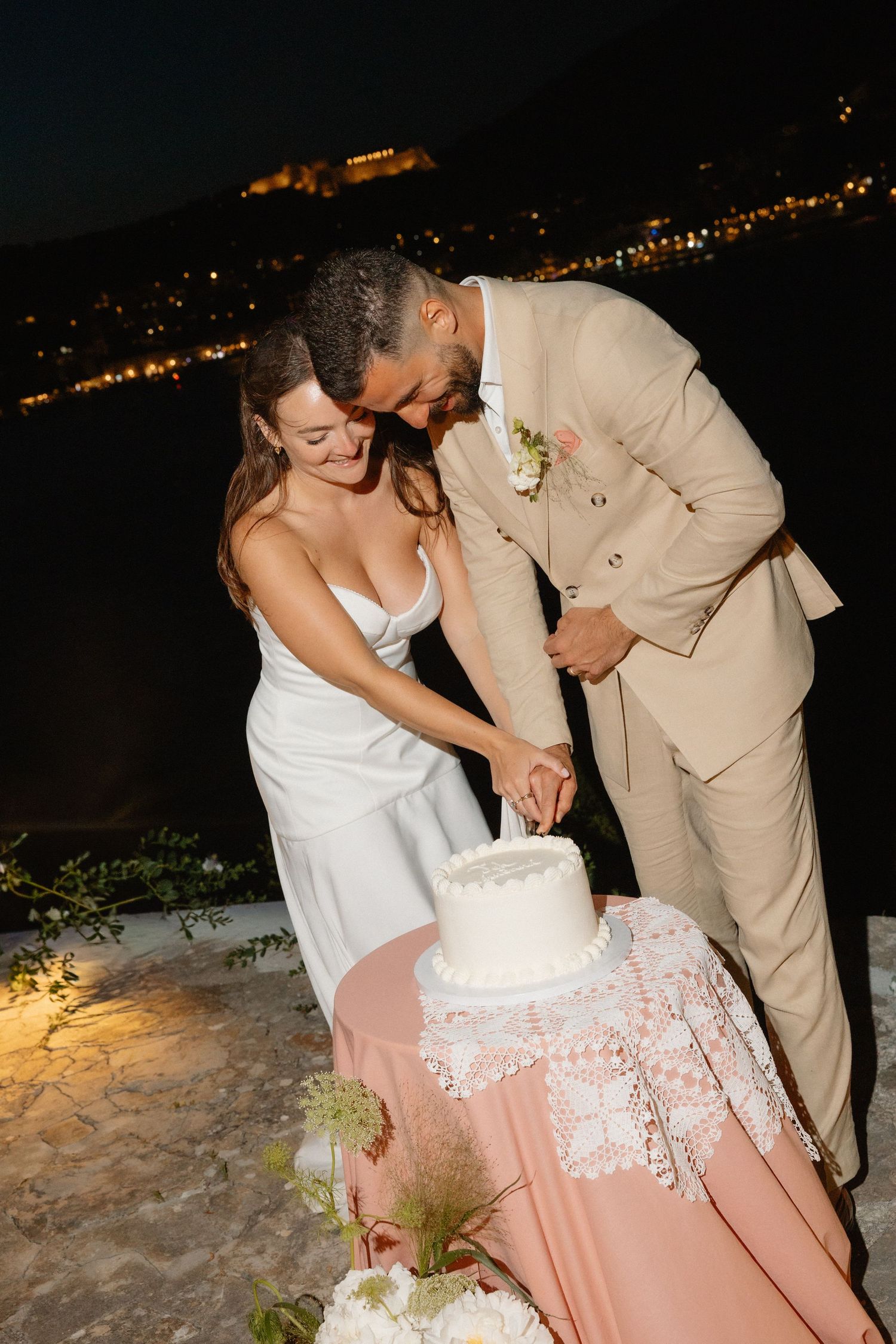 Bride and groom cutting white wedding cake together at night with city lights in the background.