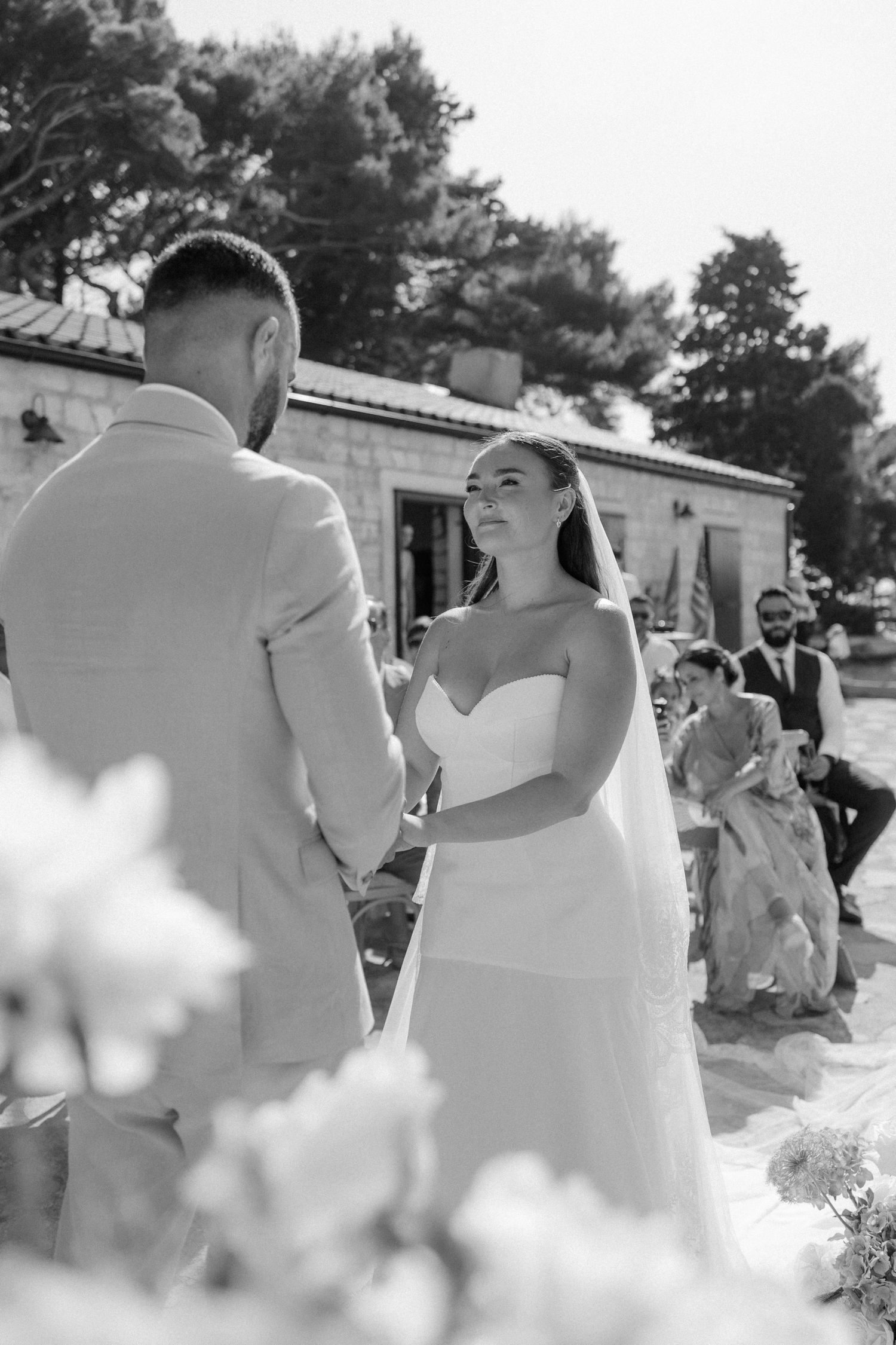 Black and white photo of a bride in a strapless gown exchanging vows with groom at an outdoor wedding ceremony.