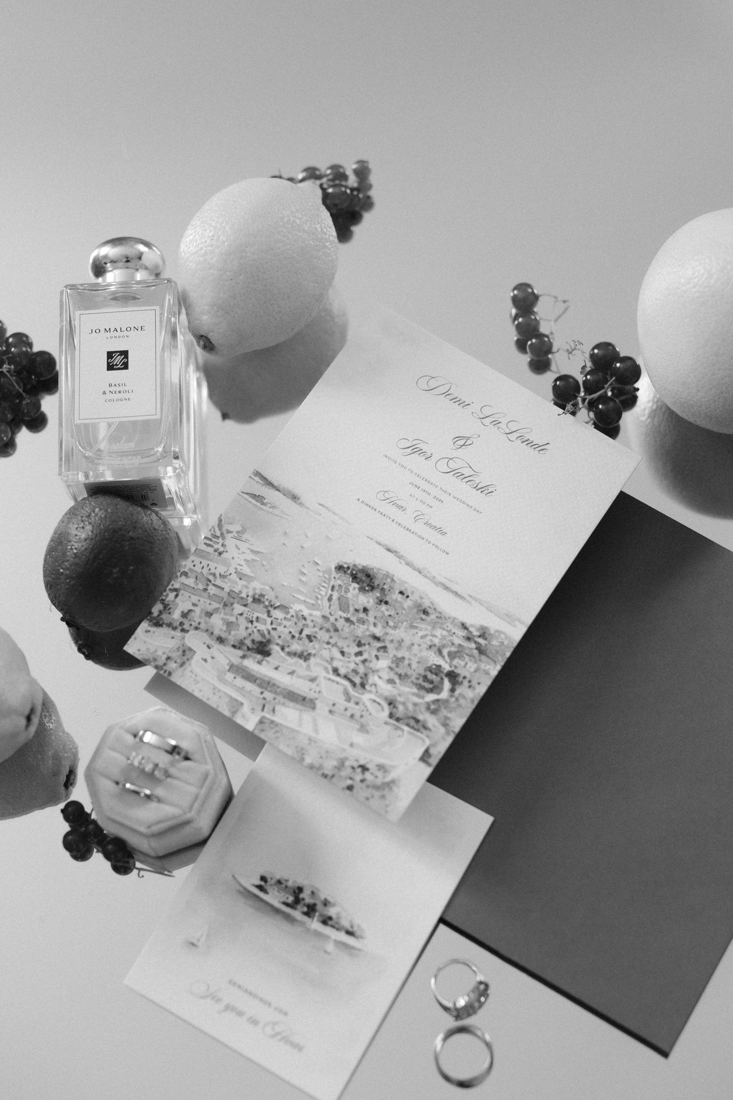Black and white flat lay of wedding invitation suite with Jo Malone perfume, rings, grapes, and fruit on light background.
