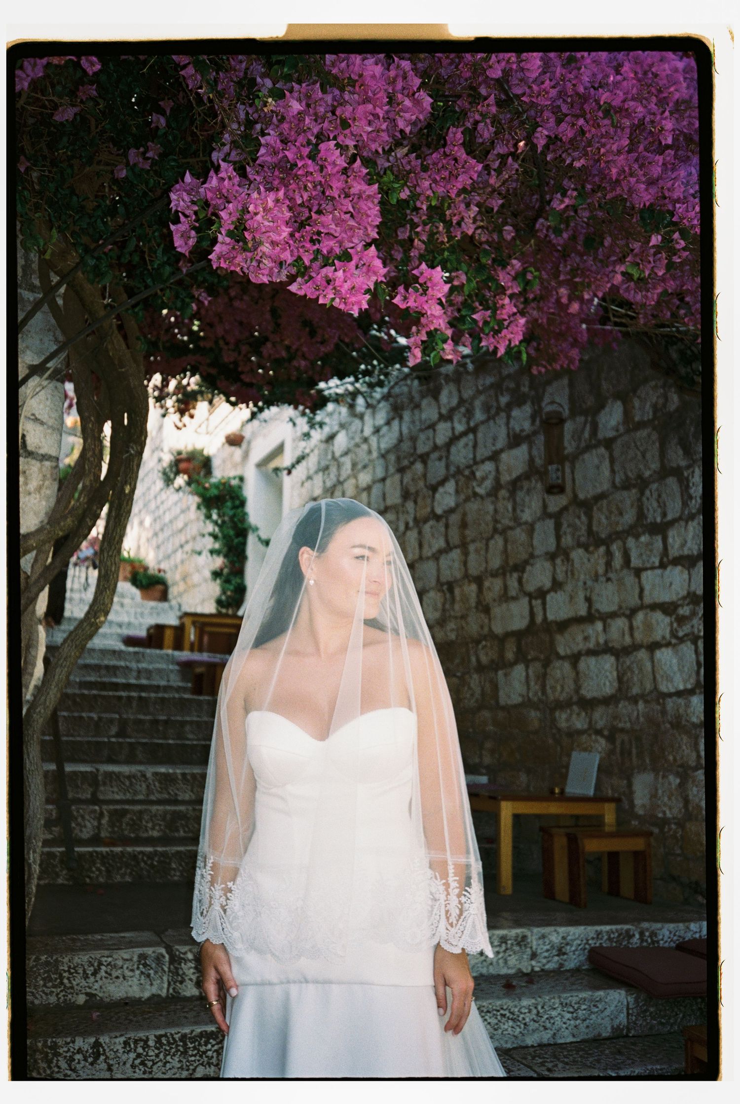 Bride in strapless gown and lace veil stands beneath vibrant pink bougainvillea.