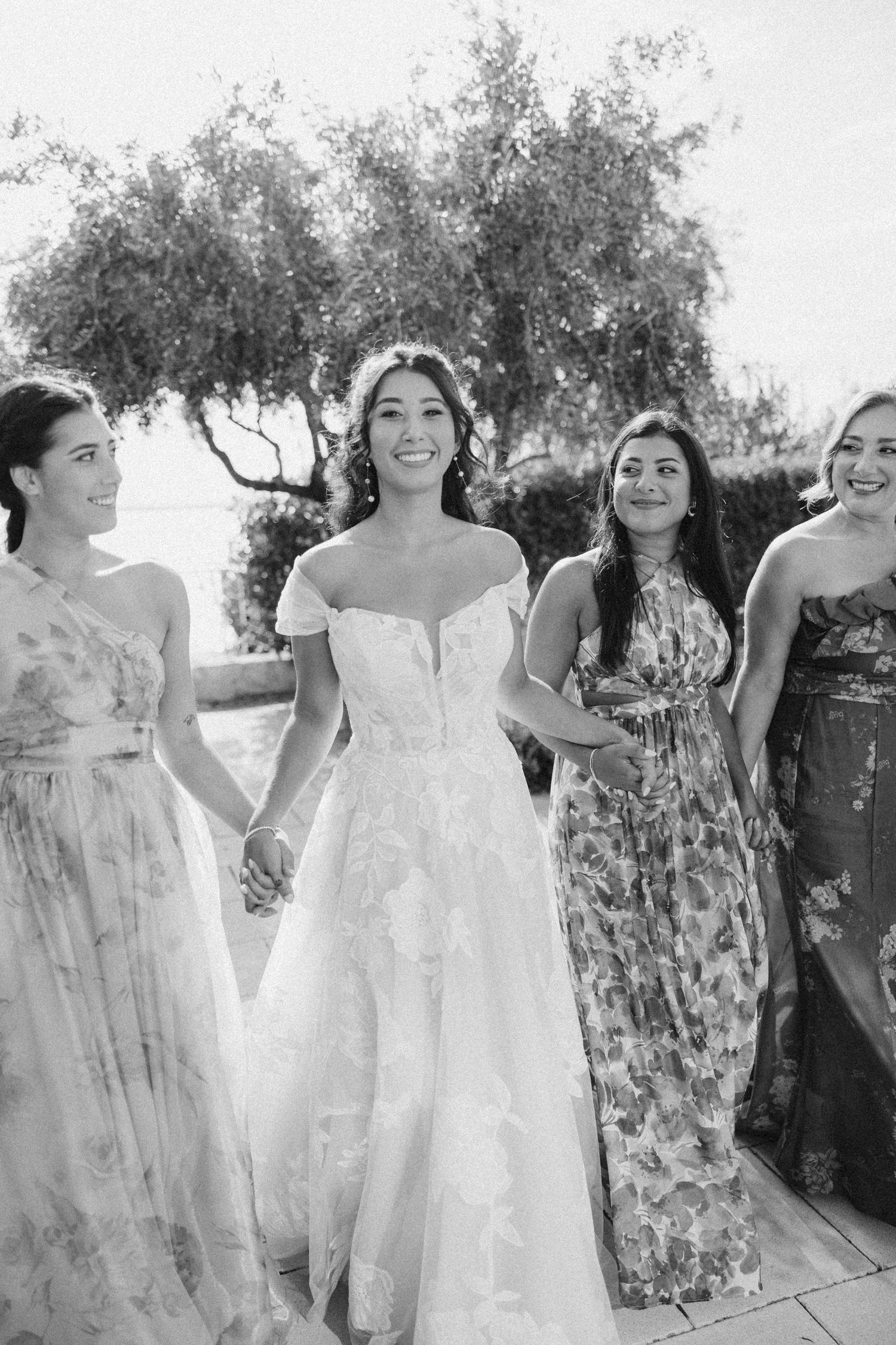 Group of bridesmaids wearing floral dresses and white gowns laughing together outdoors.