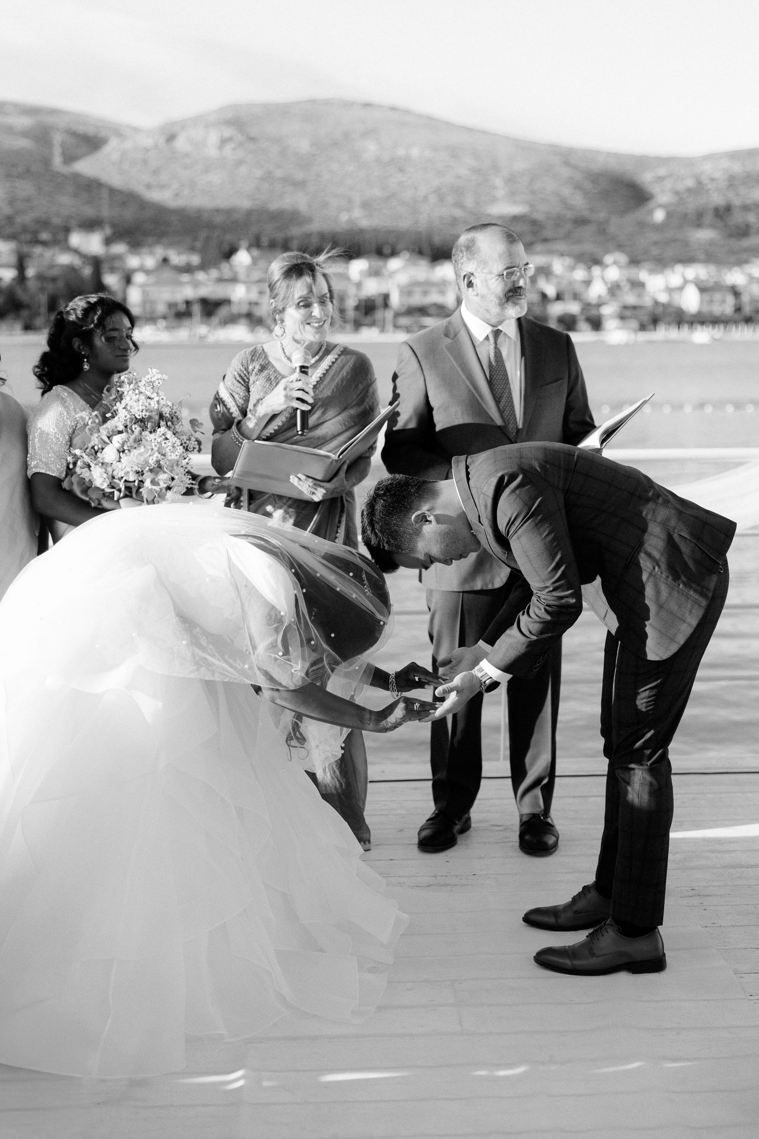 Groom bows to bride during outdoor wedding ceremony with officiant and mountains.