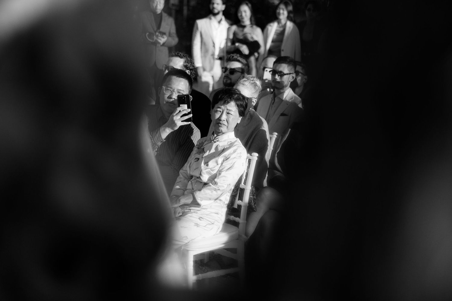 Black and white candid shot of an emotional wedding guest smiling while watching the ceremony through a crowd.