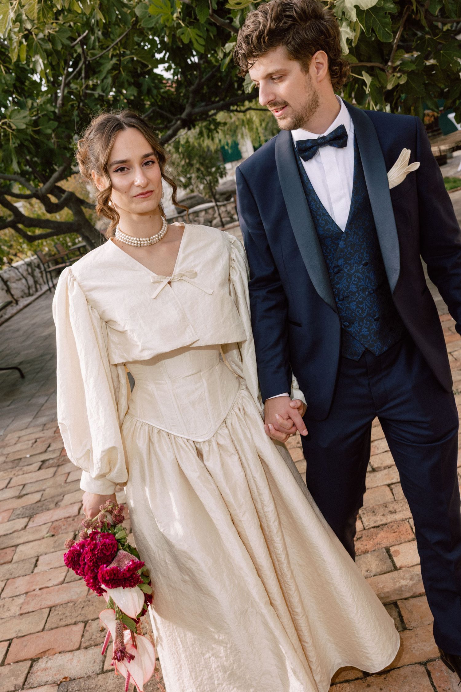 Wedding couple in vintage-inspired attire posing against brick wall.