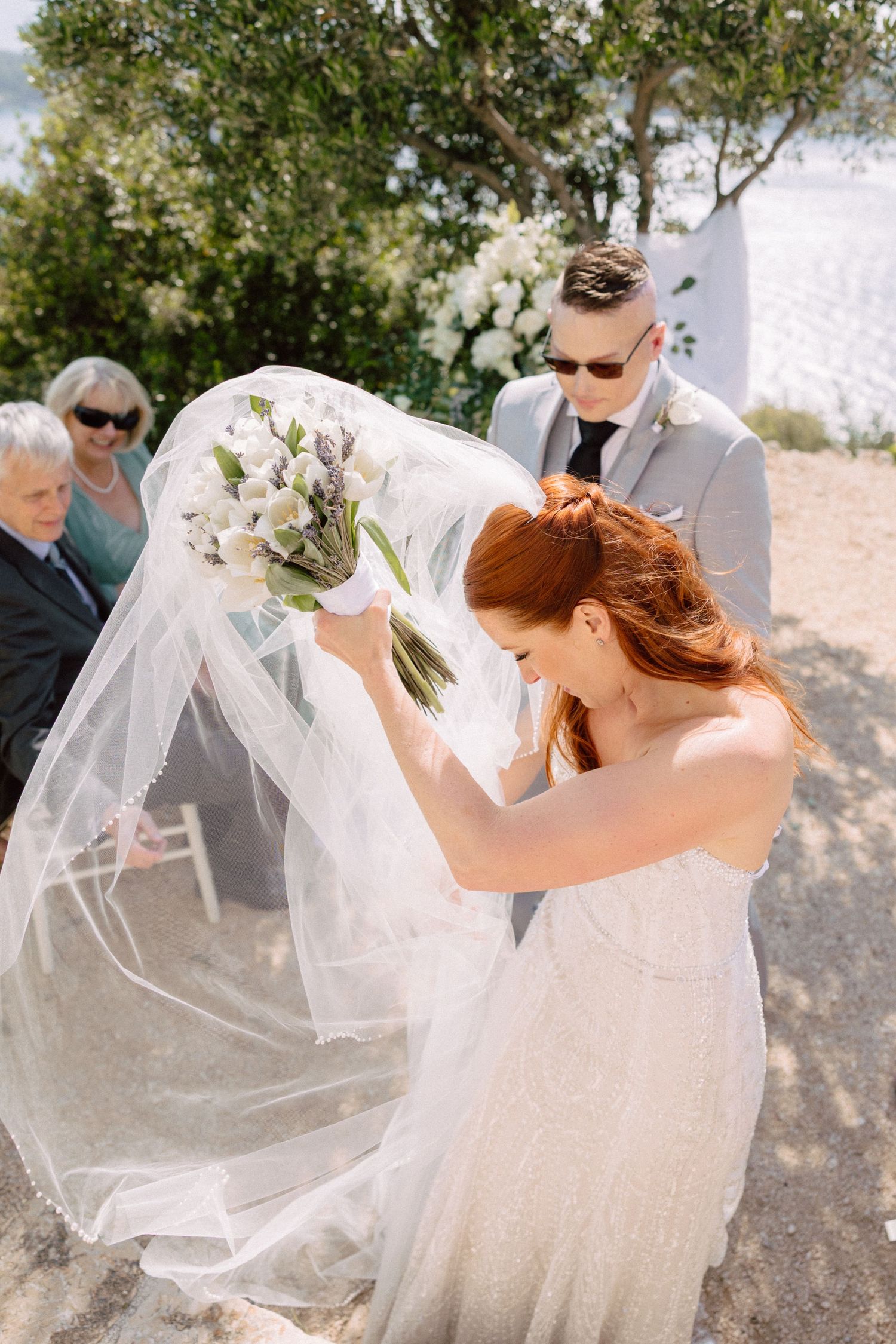 A bride's veil flows dramatically in the wind during an outdoor photoshoot.