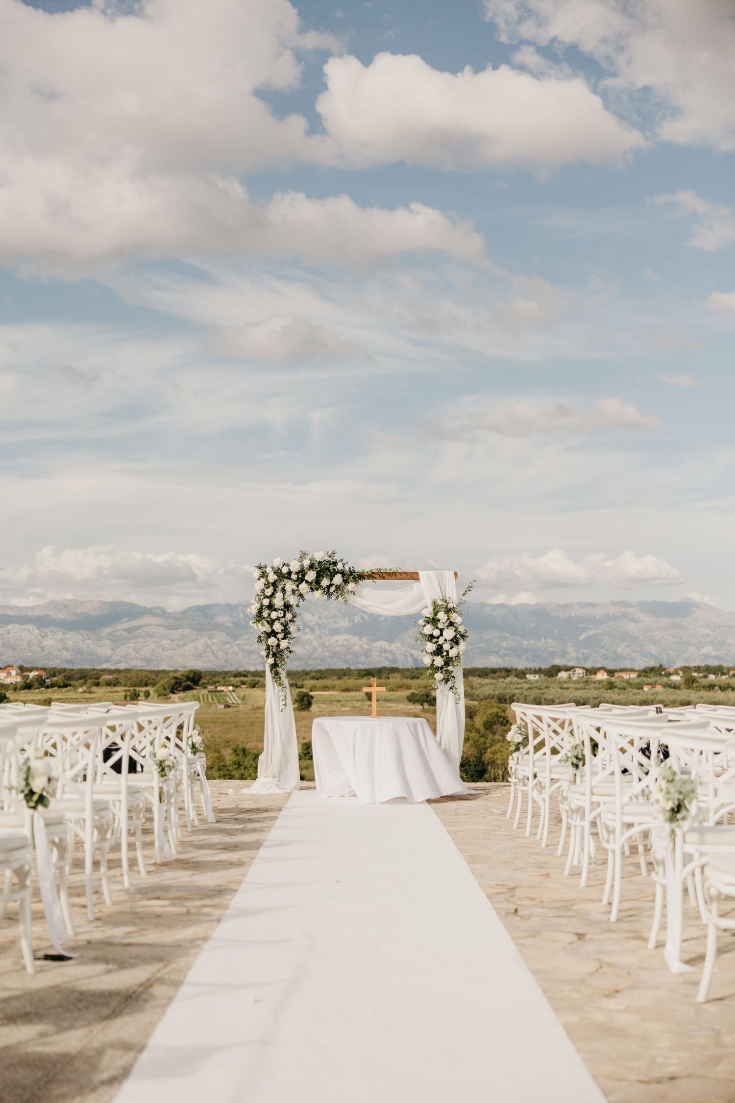 Outdoor wedding ceremony setup with white chairs and arch against cloudy mountain backdrop.