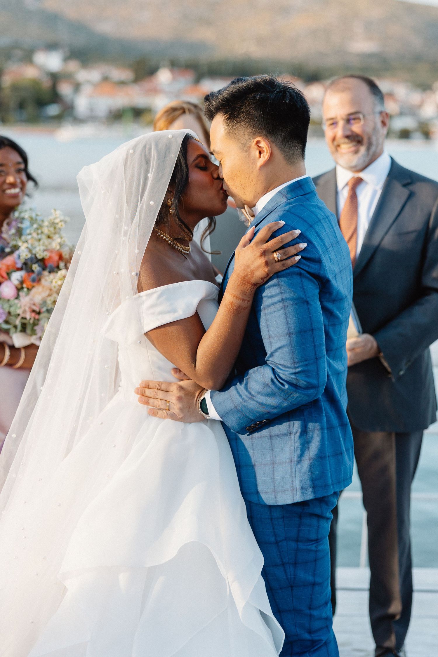 Bride and groom share first kiss at outdoor wedding ceremony, with guests smiling in background near waterfront.
