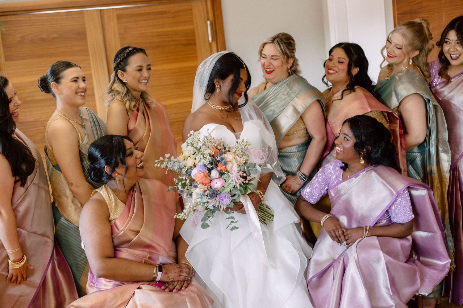 Joyful bride in white gown holding a colorful bouquet surrounded by bridesmaids in pink and sage saris.
