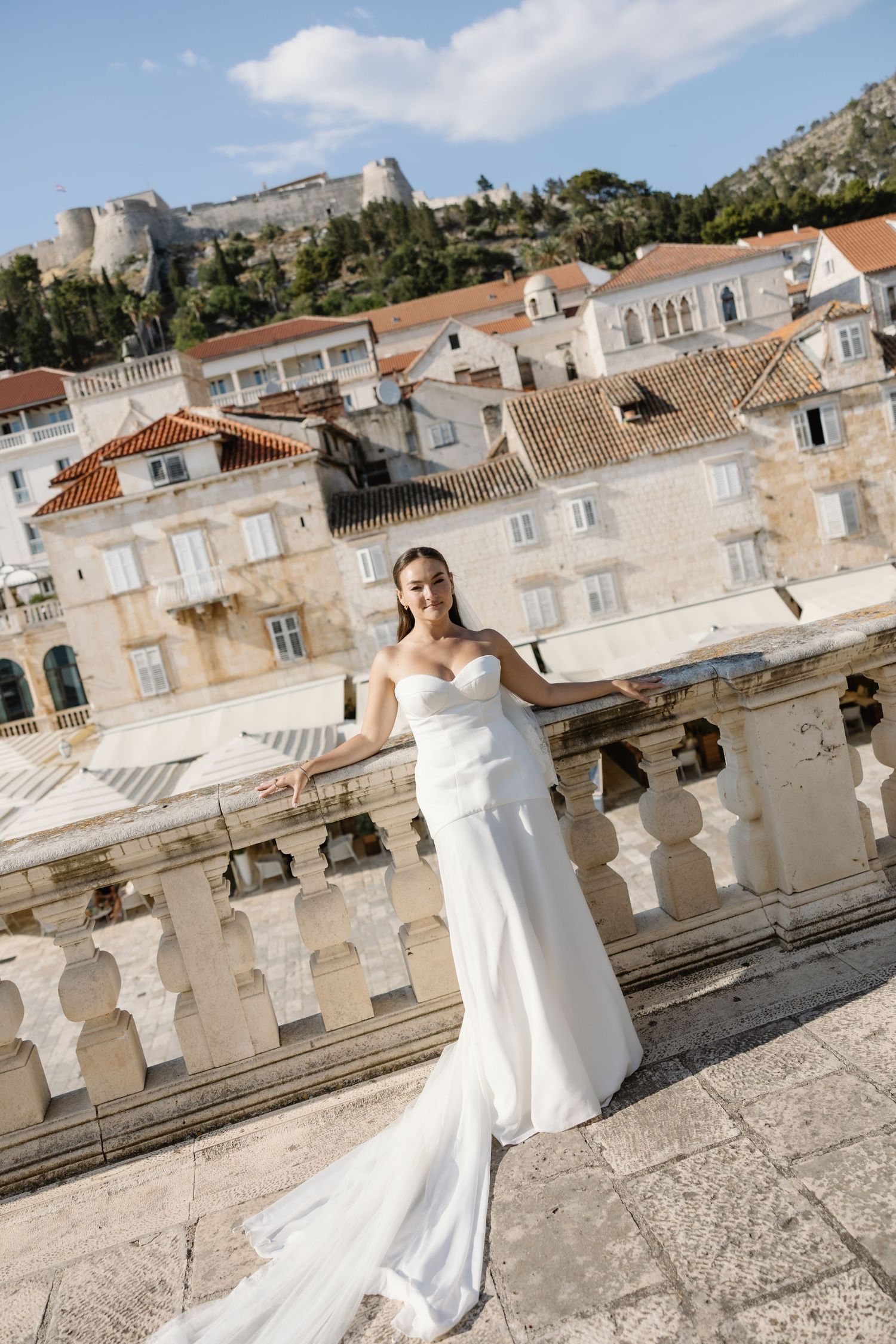 Radiant bride in white strapless gown poses on balcony with Croatian old town rooftops behind.