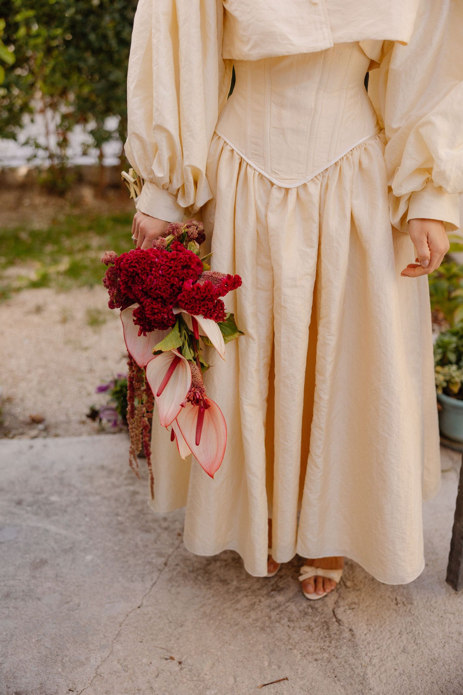 A cream colored vintage dress with puffed sleeves holds a vibrant red bouquet against a stone path.