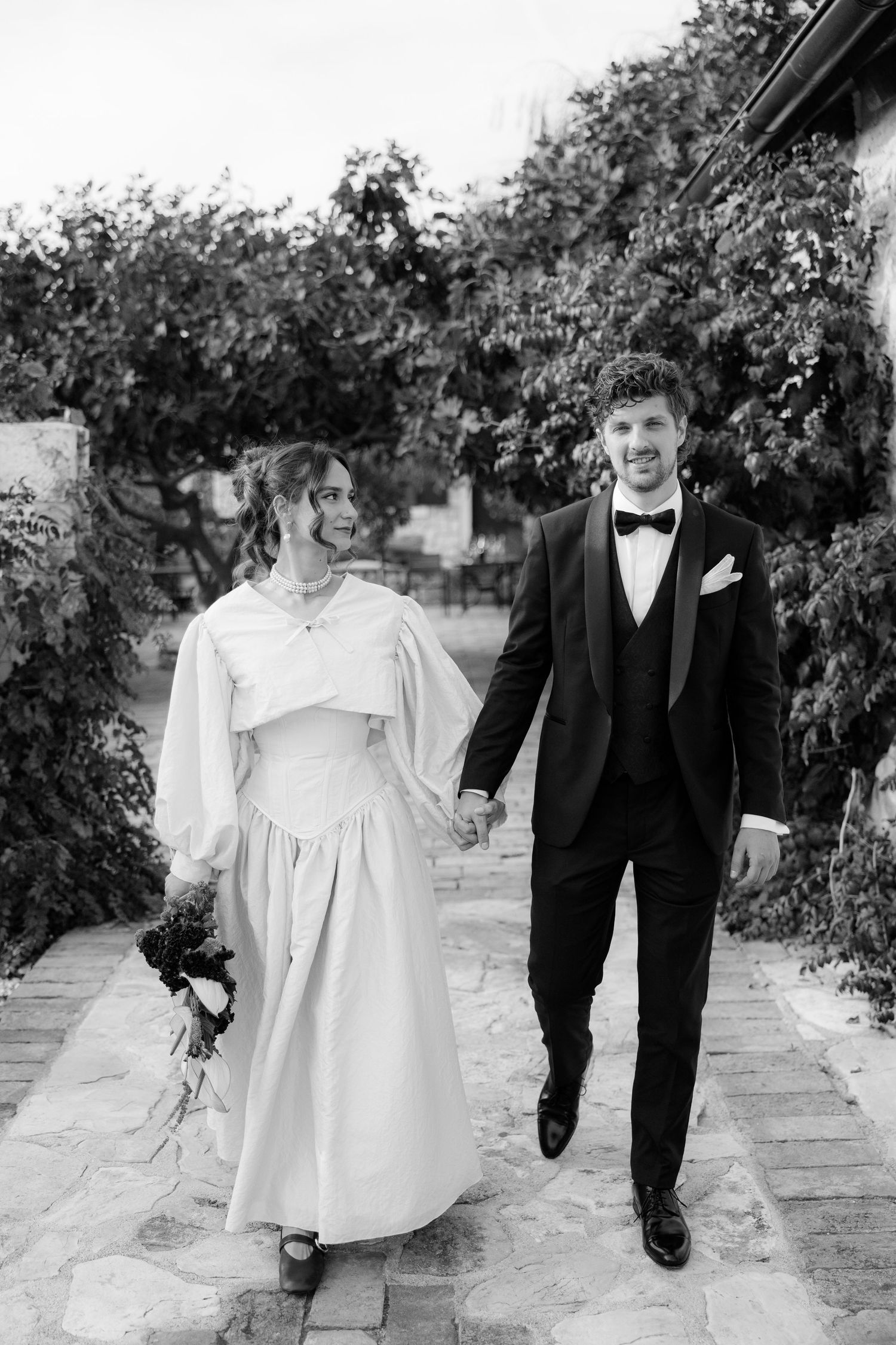 A couple walks hand in hand along a garden path in wedding attire.