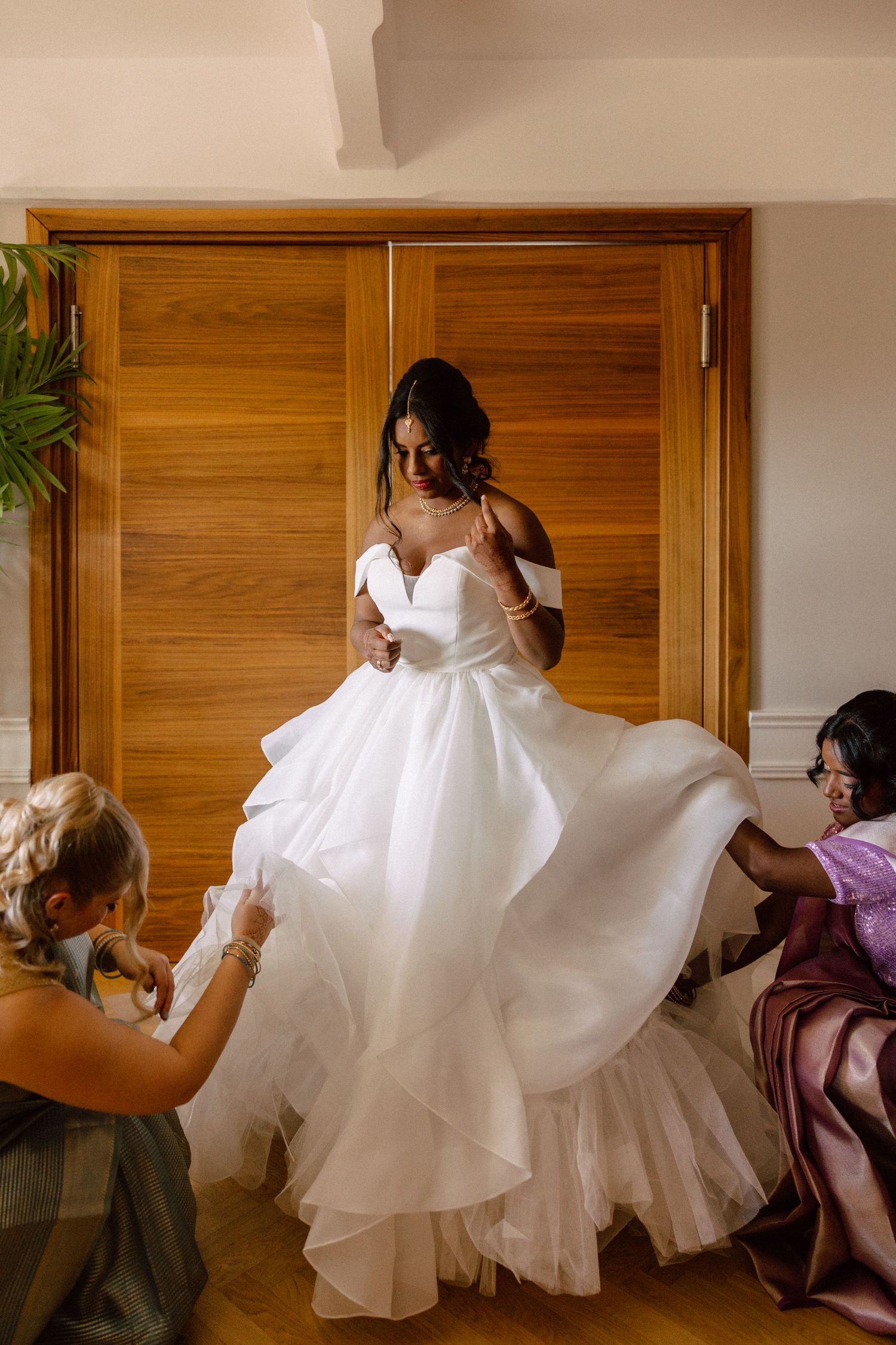 Bridesmaids helping bride into elegant off-shoulder white ball gown wedding dress with layered skirt.