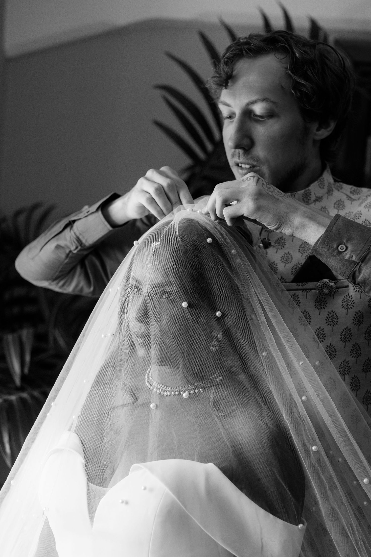 Black and white photo of a man carefully placing a pearl-embellished veil on a bride in a white wedding gown.