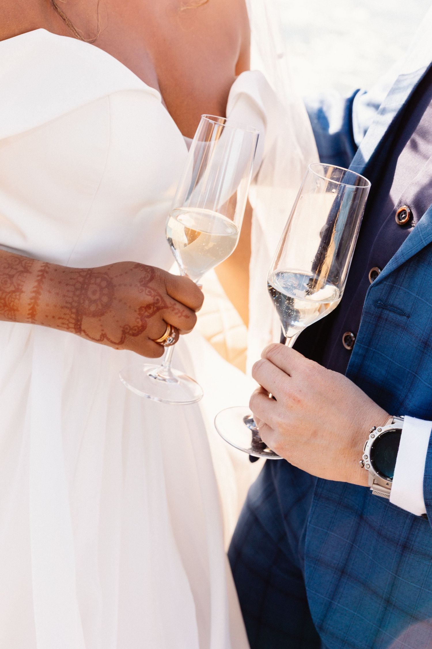 Bride and groom clinking champagne flutes at wedding, bride wearing henna and ring, groom in blue suit with watch.