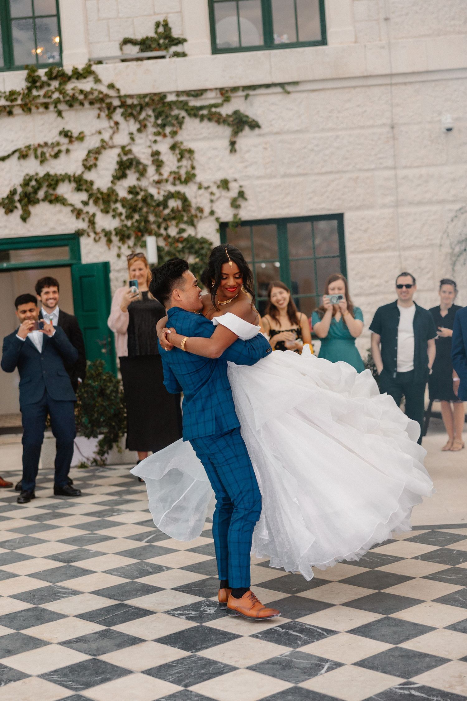 Groom lifts bride during first dance on checkered floor surrounded by applauding wedding guests outdoors.