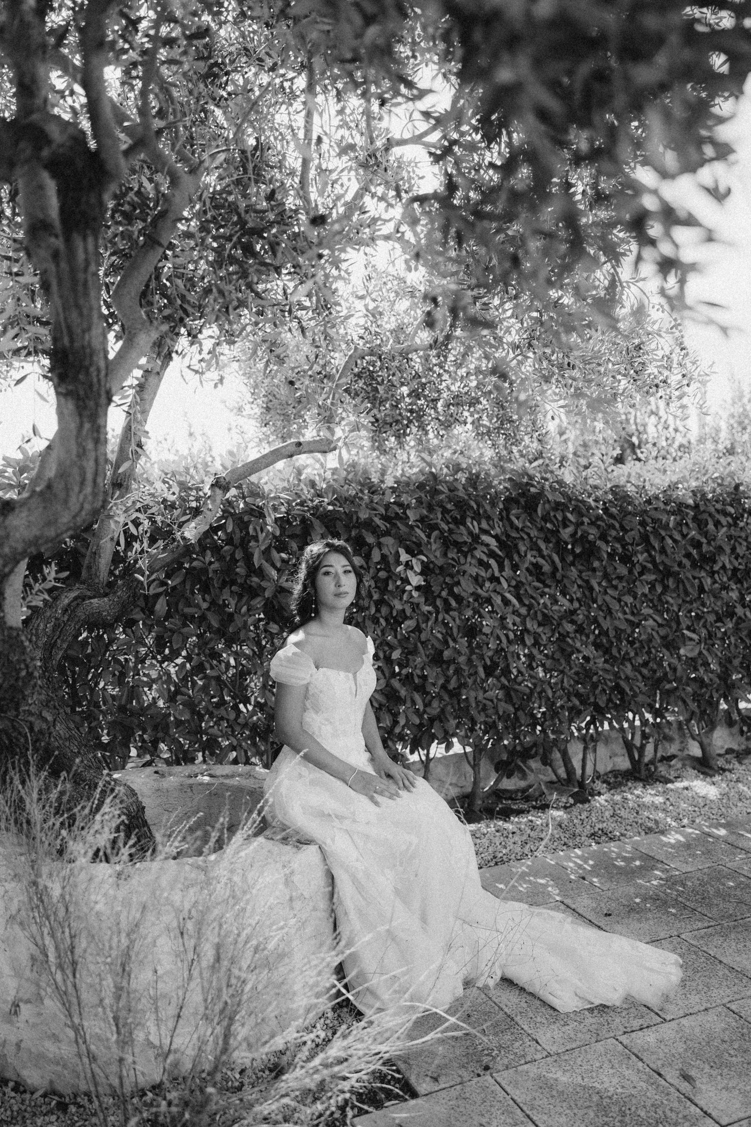 Bride in a white wedding gown posing against a garden backdrop with hedges.