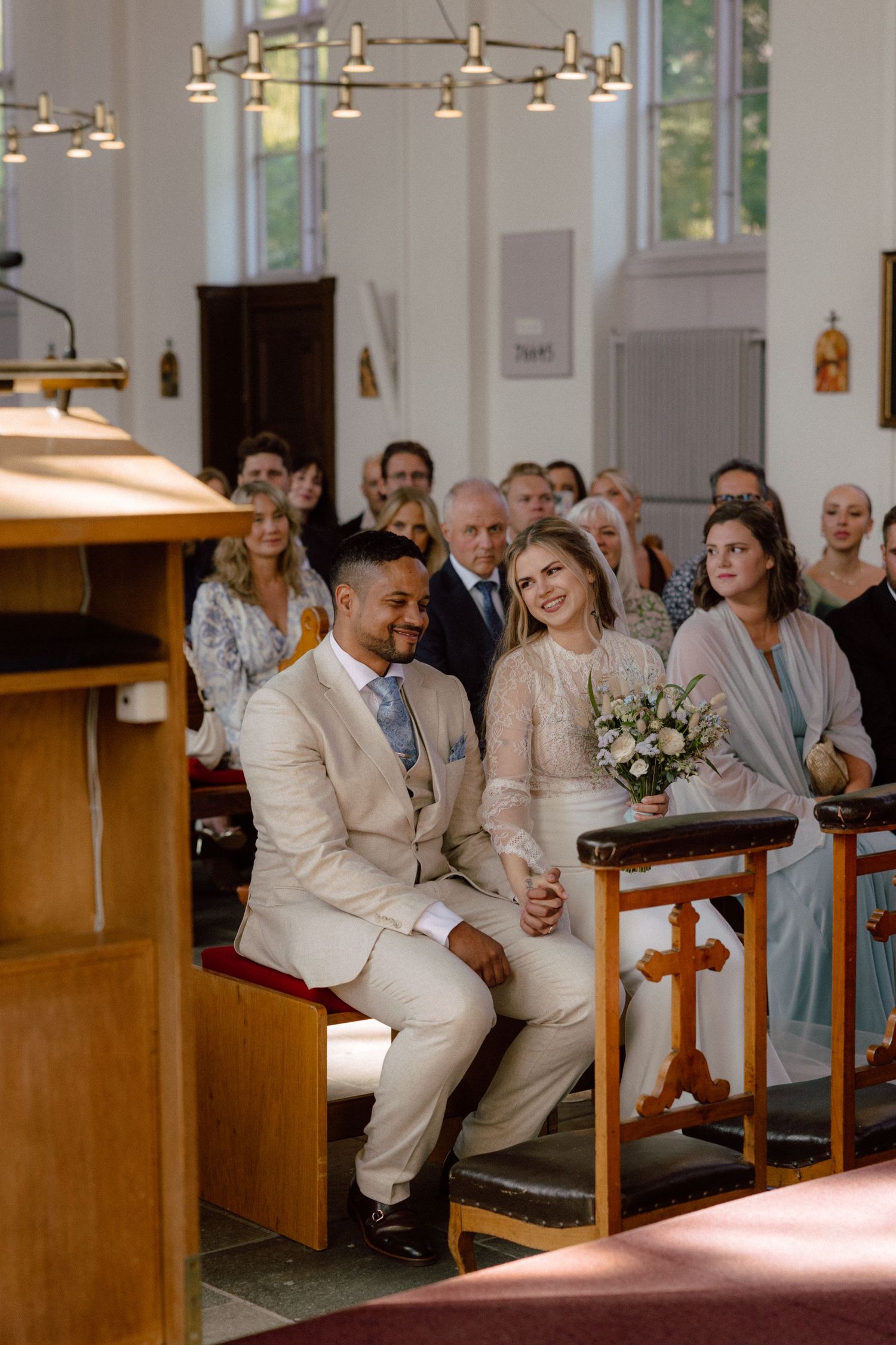Wedding party members sit together in light-filled church pews during the ceremony.