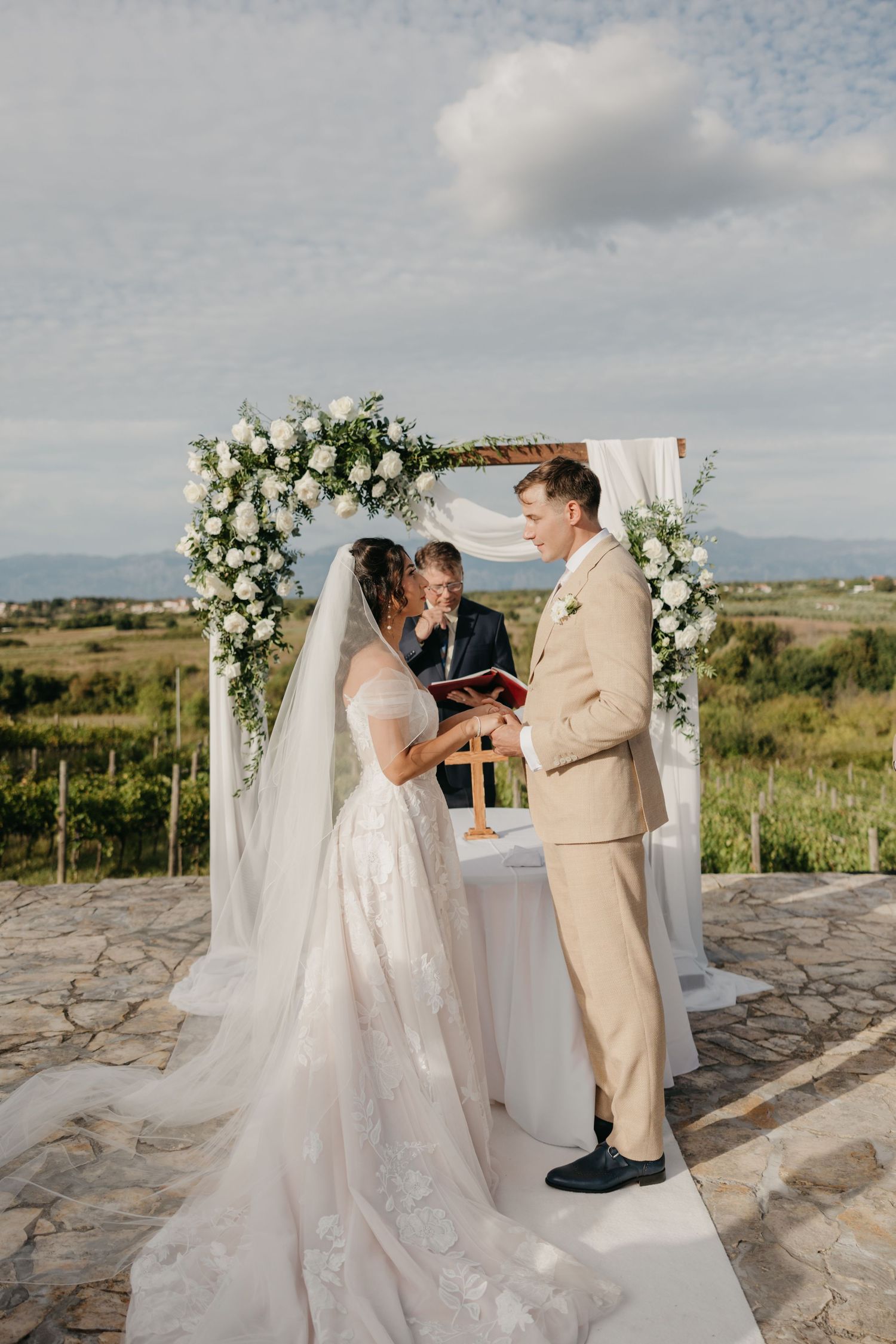 A couple exchanges vows under a floral arch at an outdoor wedding ceremony with mountain views in the background in Italy.