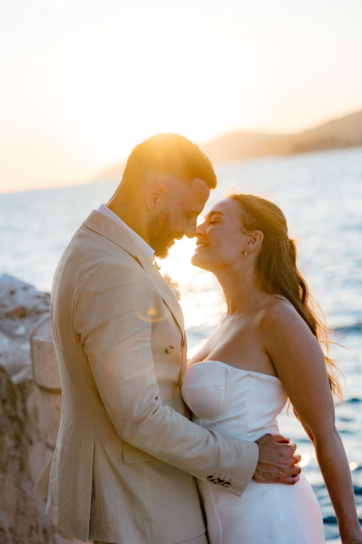 Romantic couple nearly kissing backlit by golden sunset over the ocean on their wedding day.