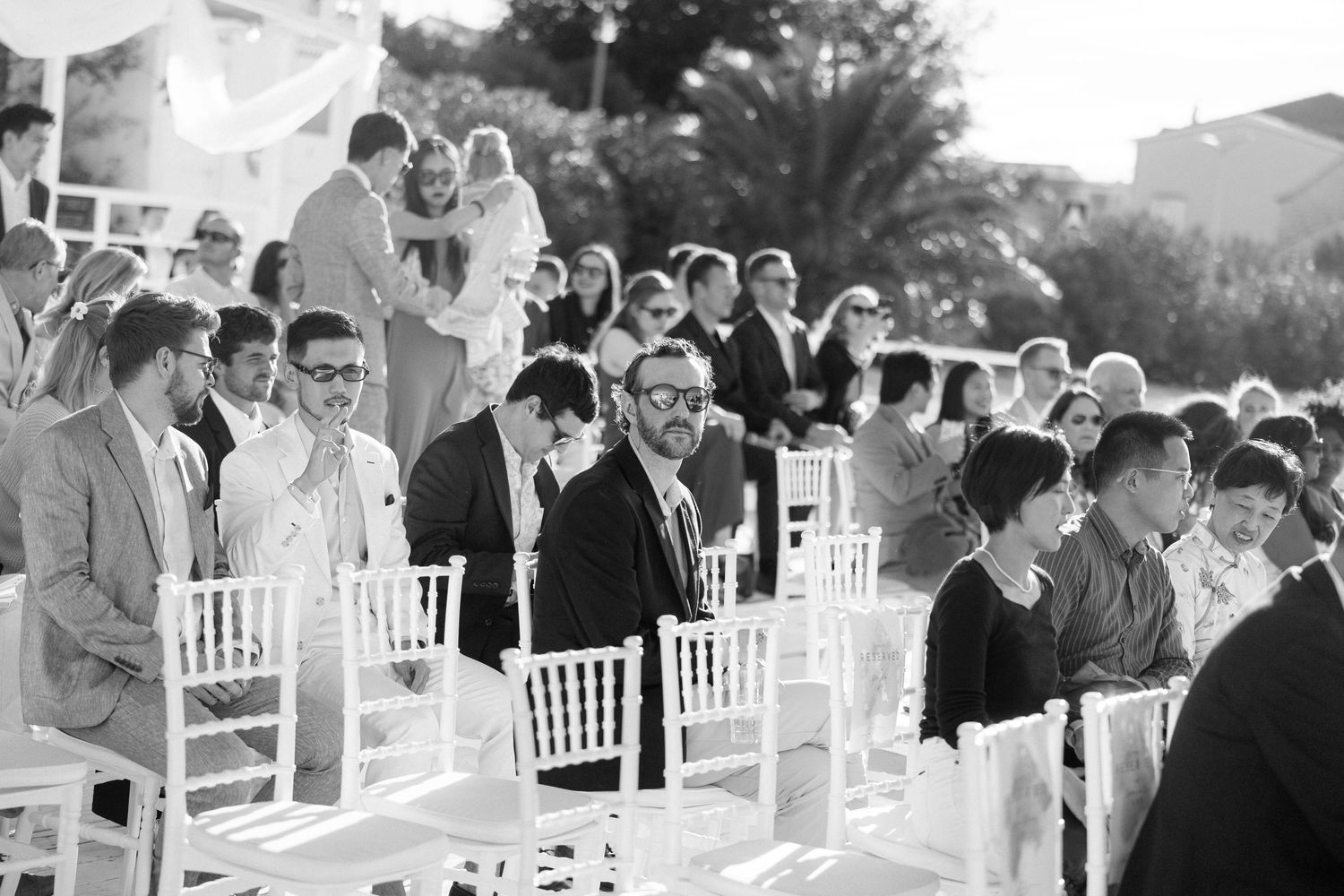Black and white photo of wedding guests seated in white chairs at outdoor ceremony.