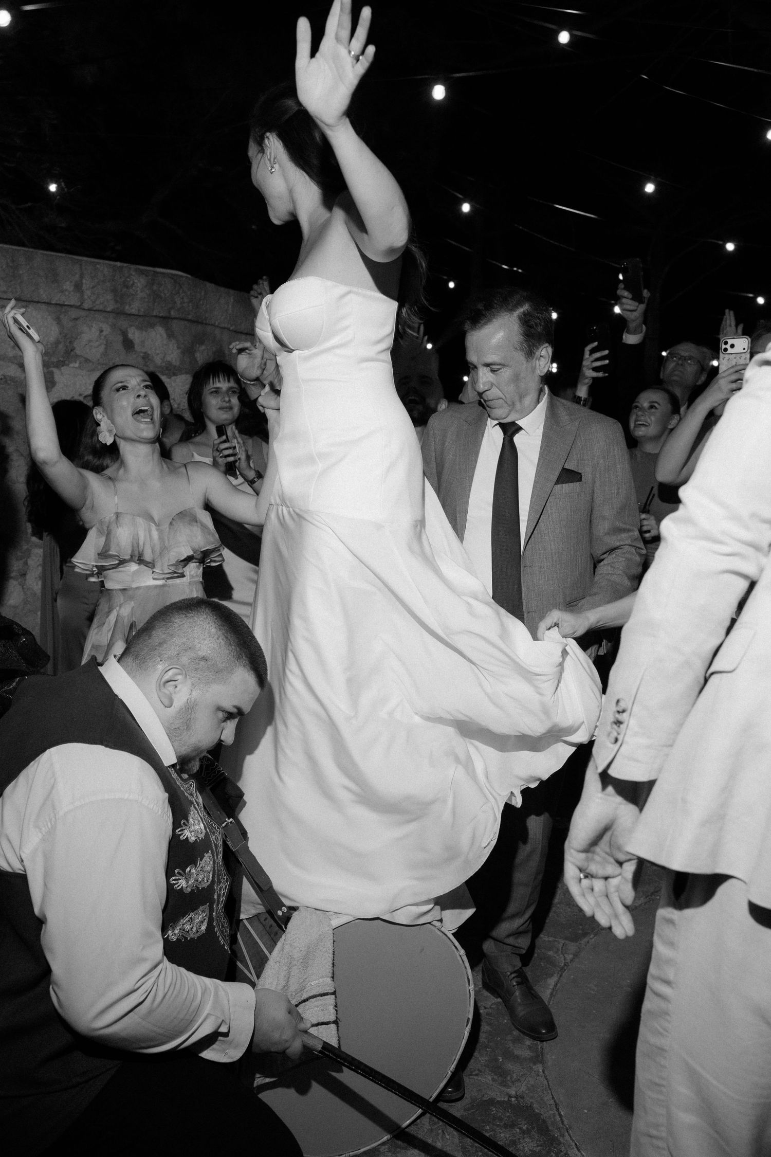Black and white photo of bride being lifted on chair during lively wedding reception celebration.