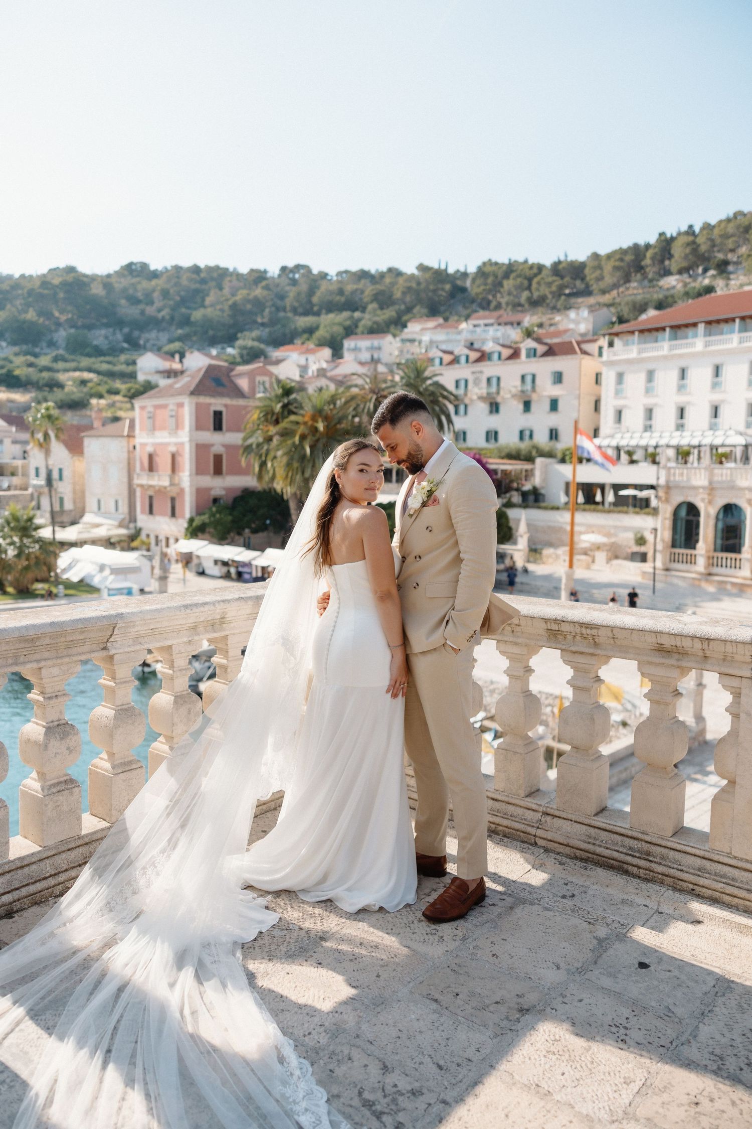 Bride and groom embracing on ornate stone balcony with colorful Croatian coastal town buildings in background.