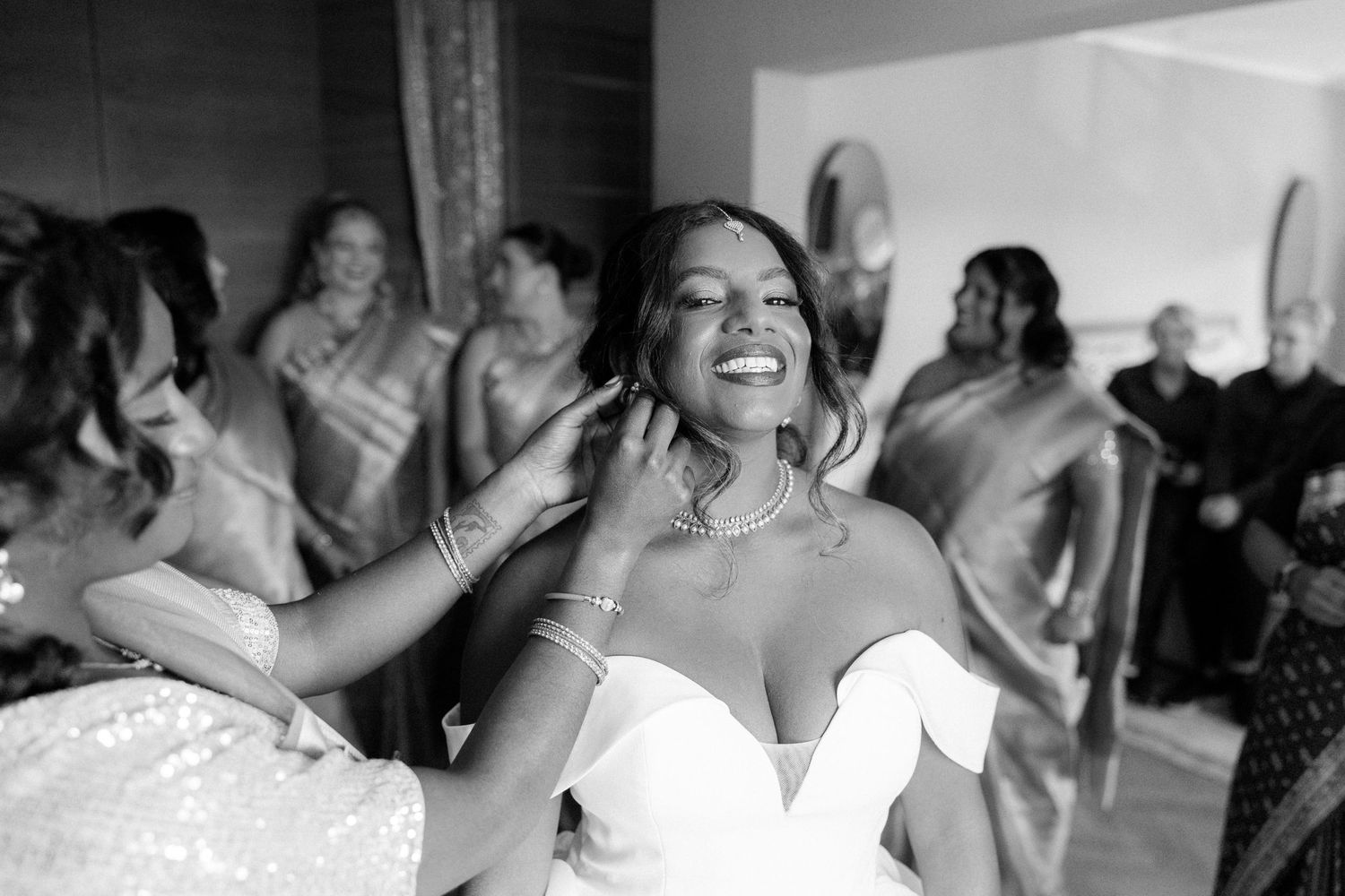 Black and white photo of a joyful bride smiling as bridesmaids help her prepare, with guests in saris watching.