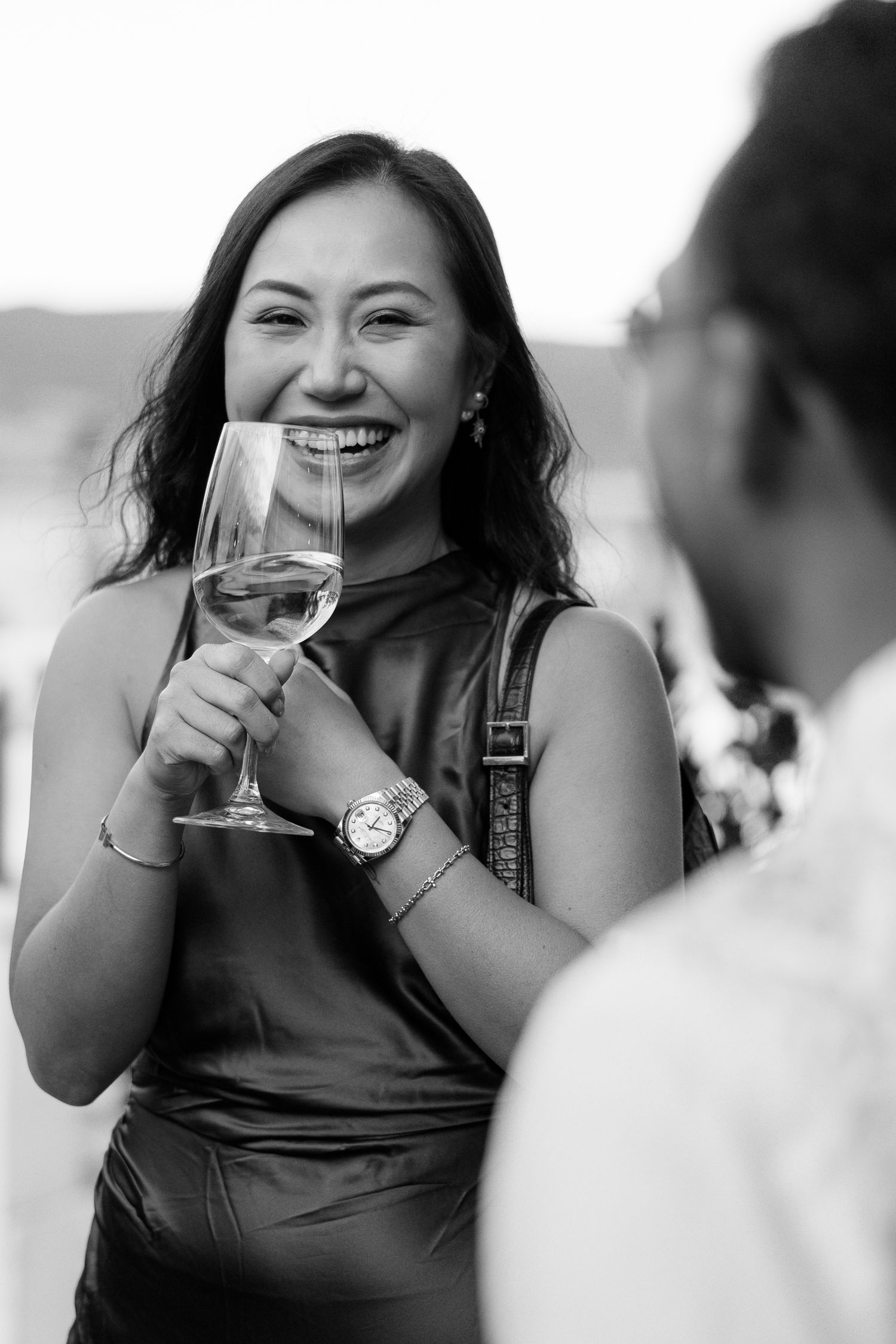 Smiling Asian woman holds a wine glass at an outdoor wedding reception in black and white.