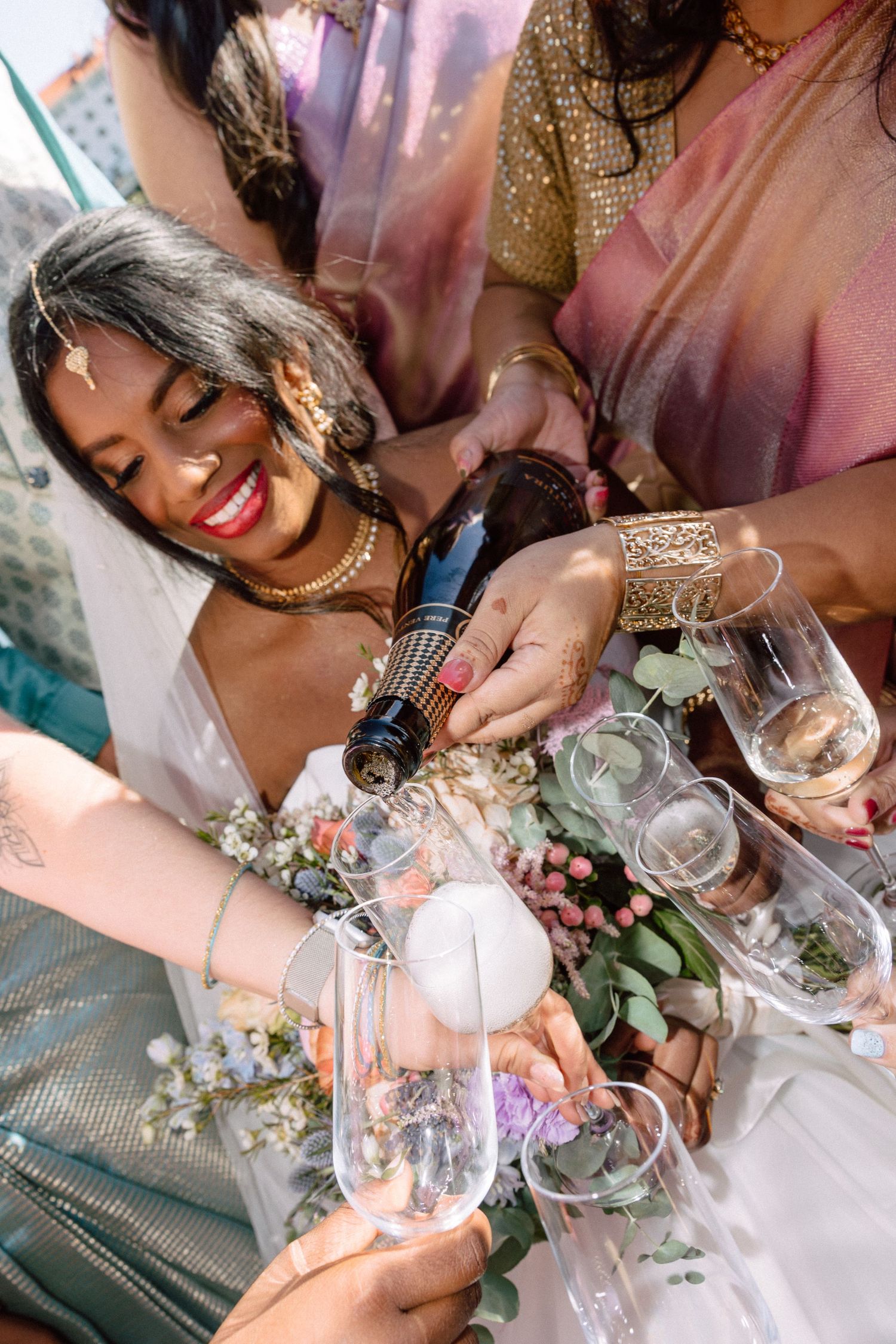 Smiling bride in Indian attire pouring champagne with bridesmaids in colorful sarees.