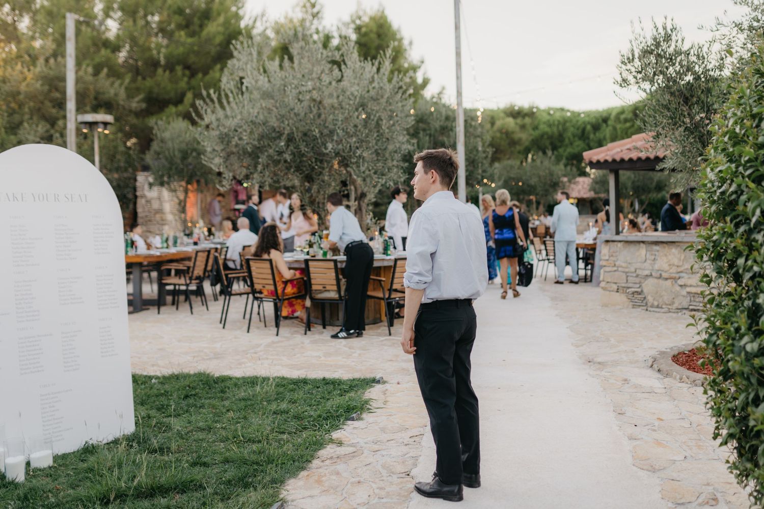 Wedding guests walking along stone pathway lined with olive trees.