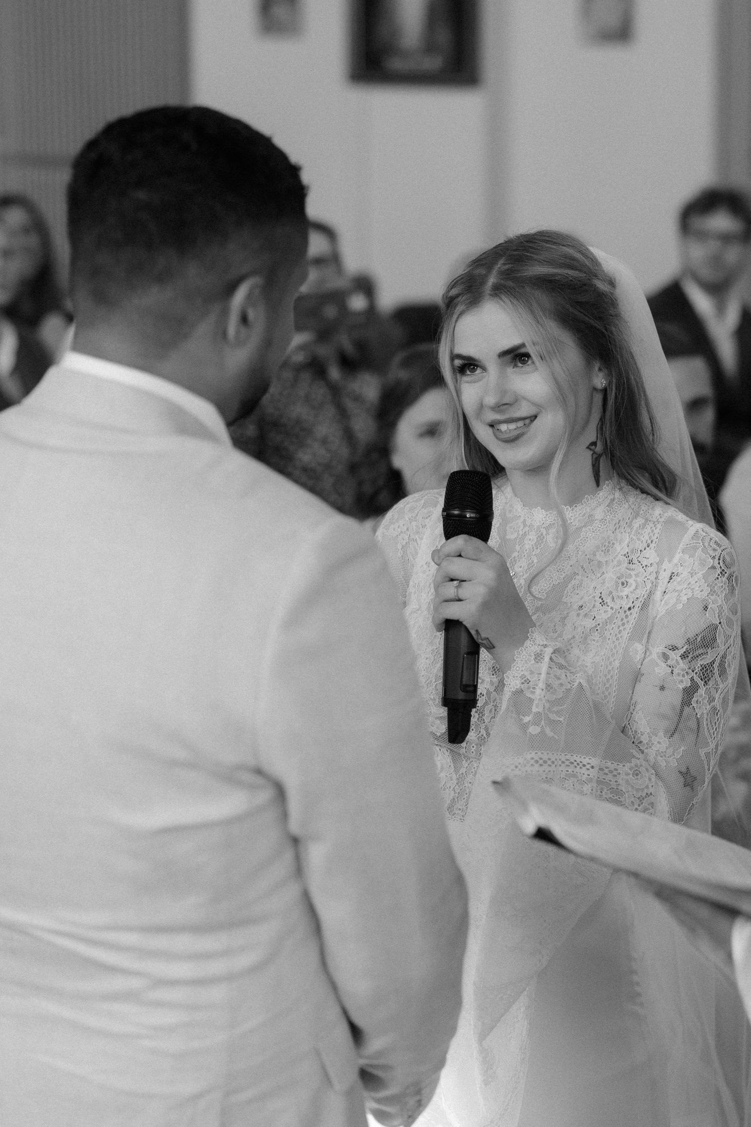 Bride in lace dress smiles while holding microphone during wedding ceremony in black and white.