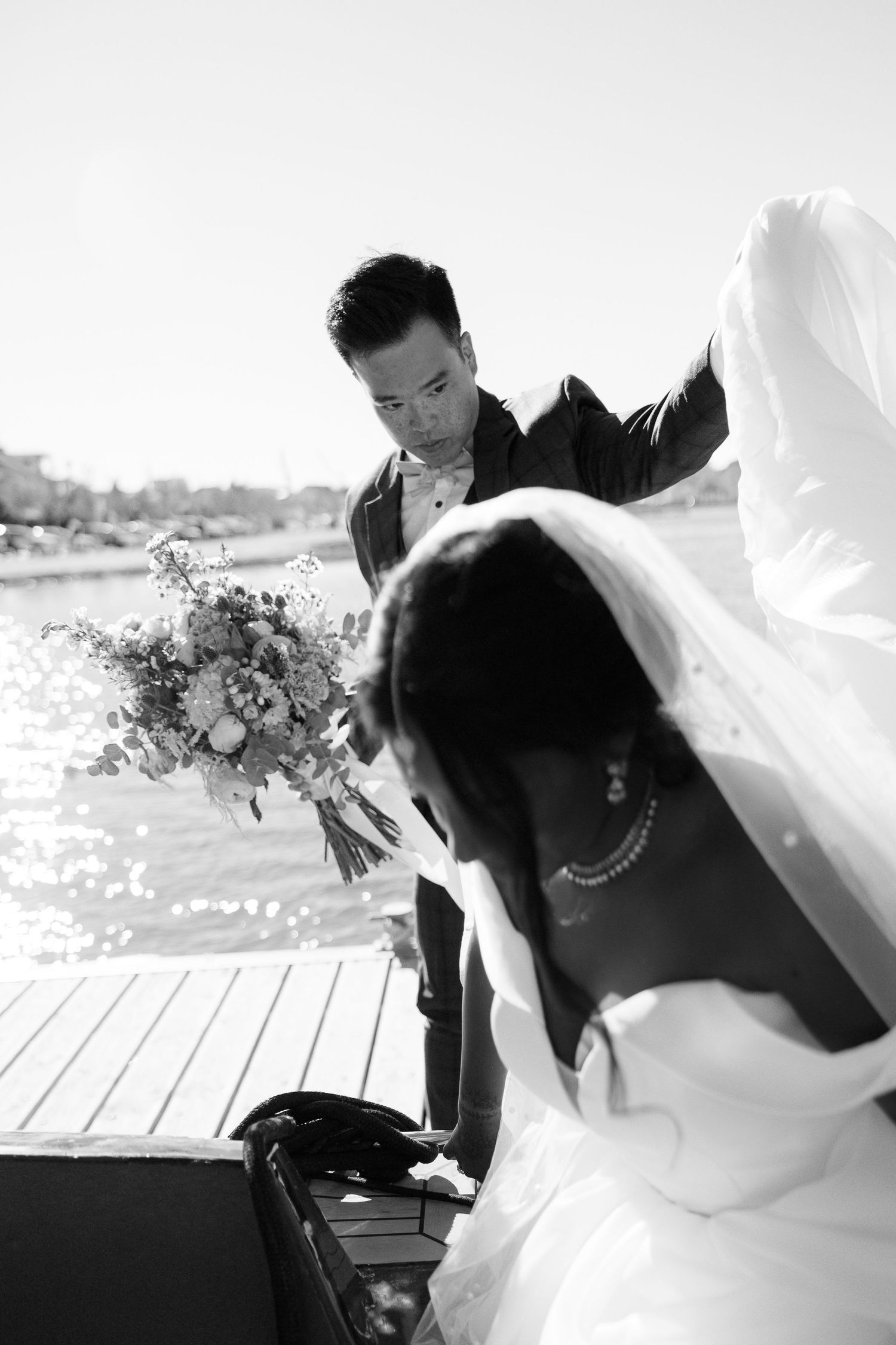 Groom hands bouquet to bride in white gown and veil on waterfront dock in black and white photo.