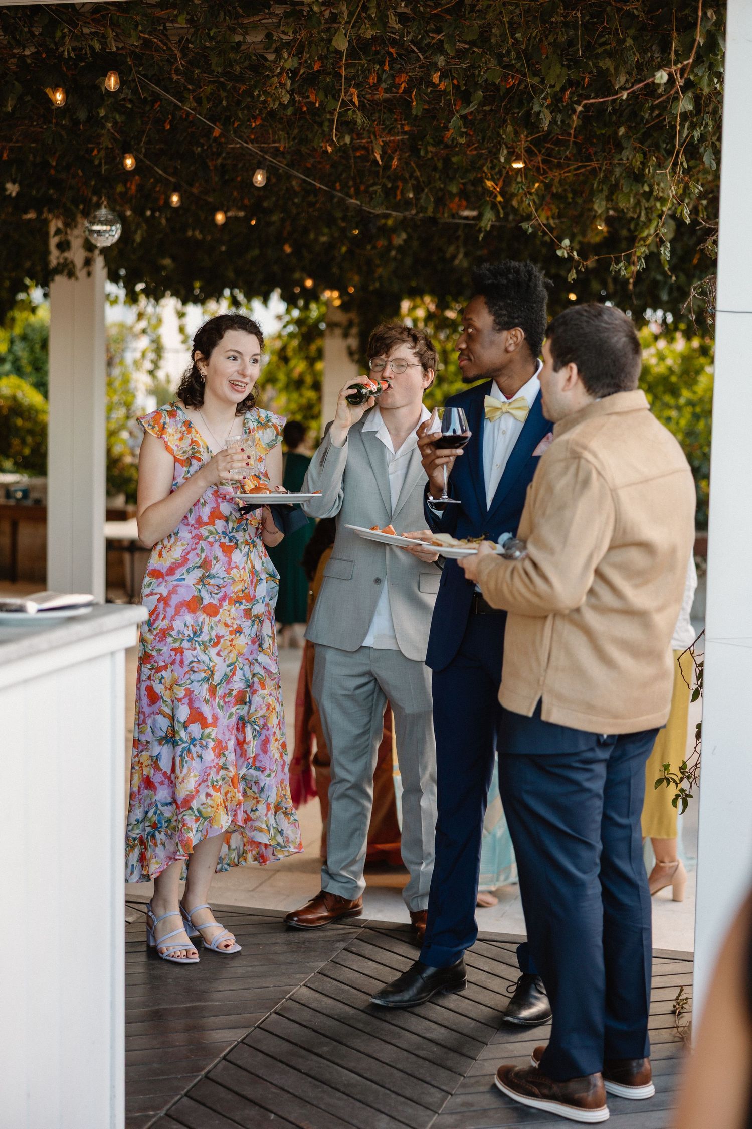 Diverse group of wedding guests socialize with wine and appetizers on decorated outdoor terrace.