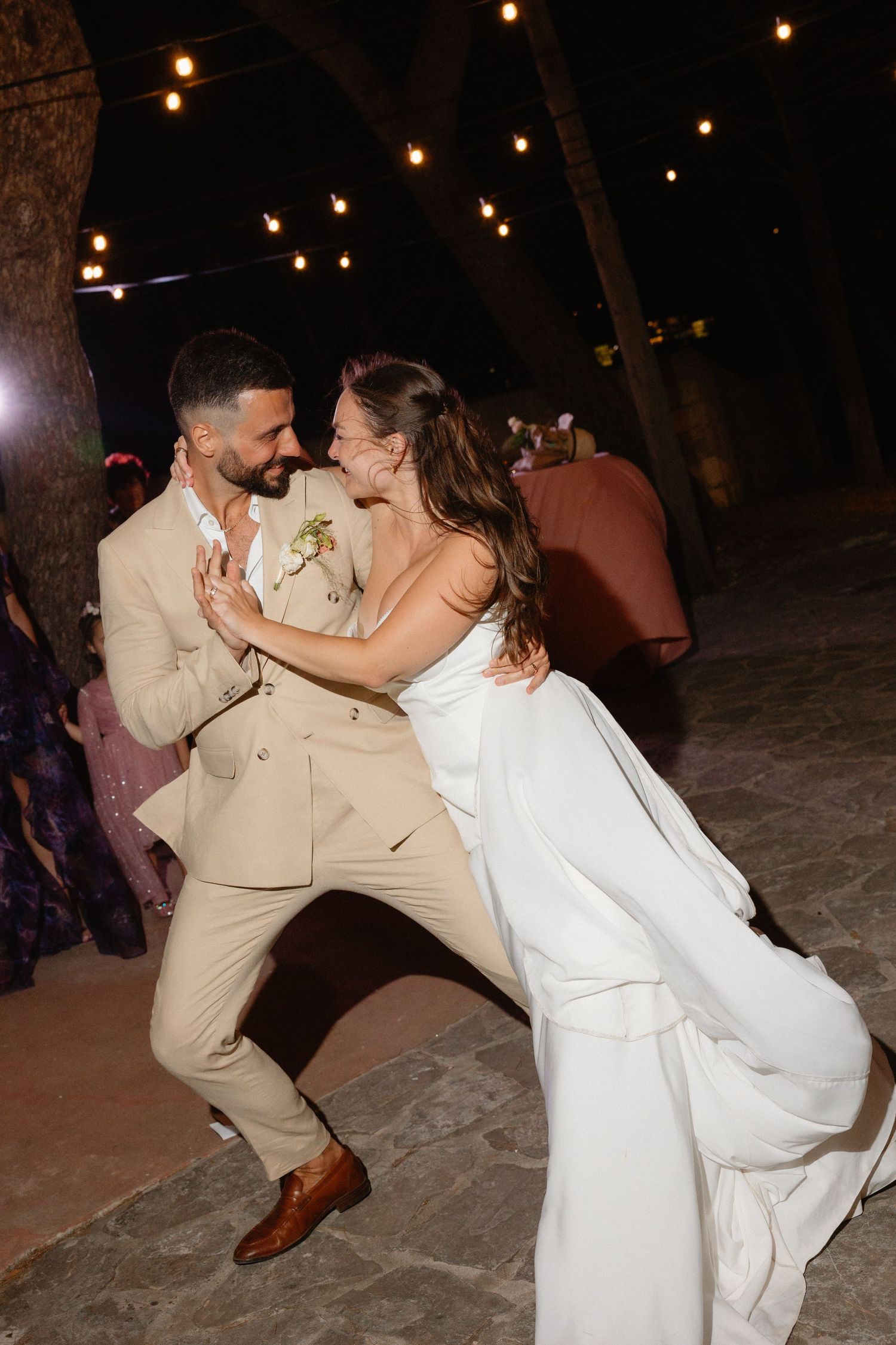 Bride and groom laugh joyfully while dancing under string lights at outdoor wedding reception.