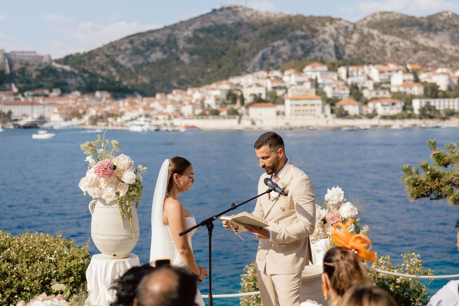 Couple exchanges vows at outdoor wedding ceremony overlooking Dubrovnik coastline with floral arrangements and mountain backdrop.