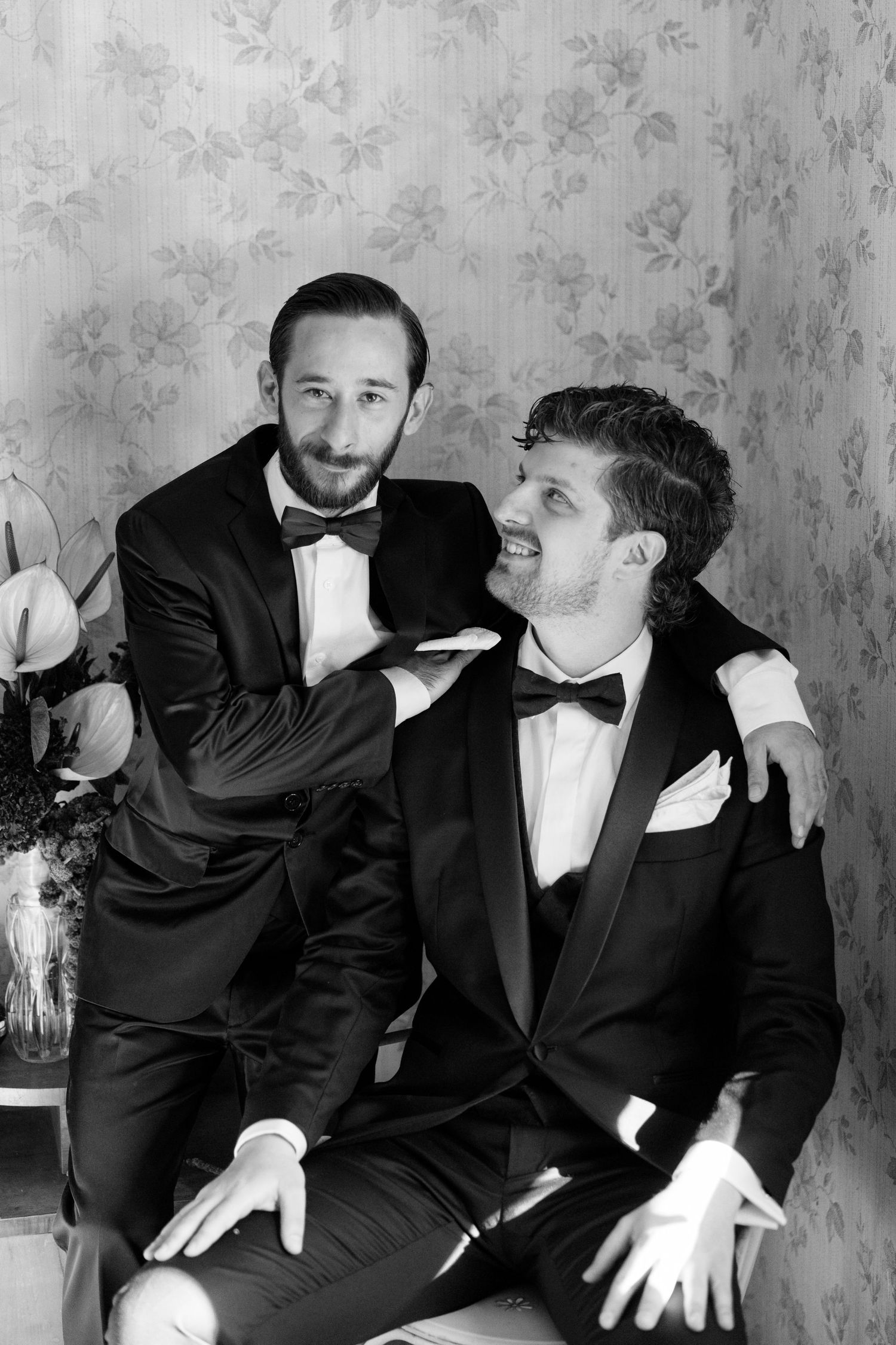 Two grooms in black tuxedos share a candid moment against a vintage floral wallpaper.