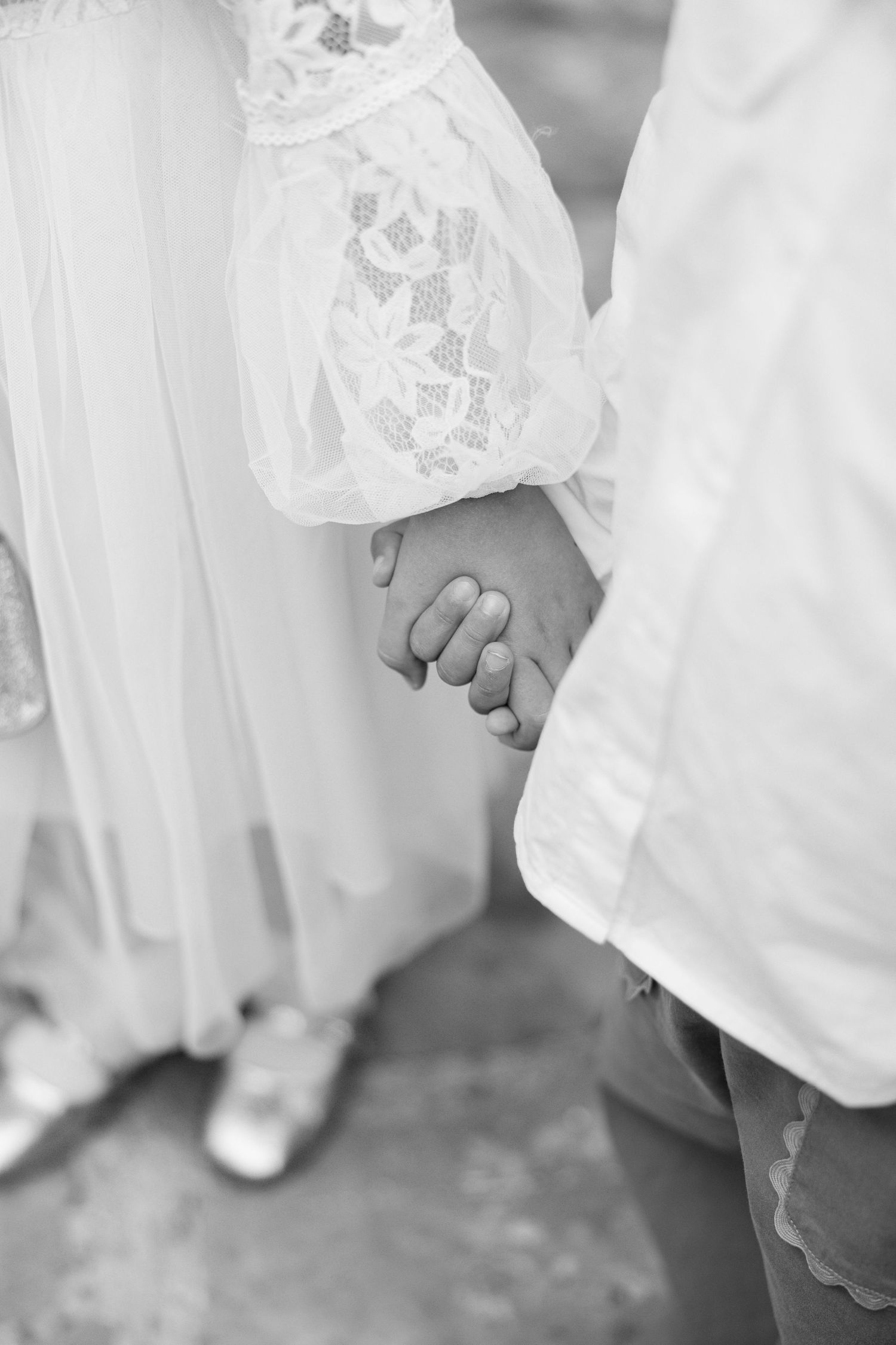 A black and white close-up of newlyweds holding hands, showing lace dress details.