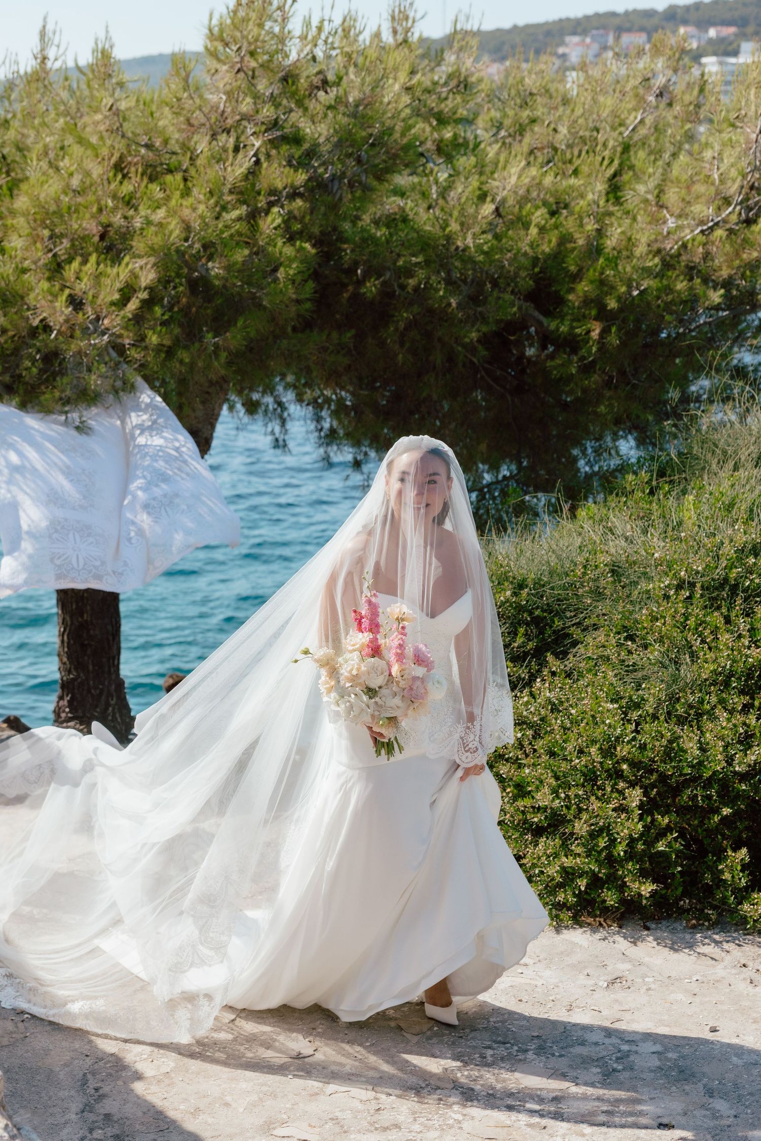 Bride in flowing white gown and veil holding bouquet walks along waterfront surrounded by lush green trees.