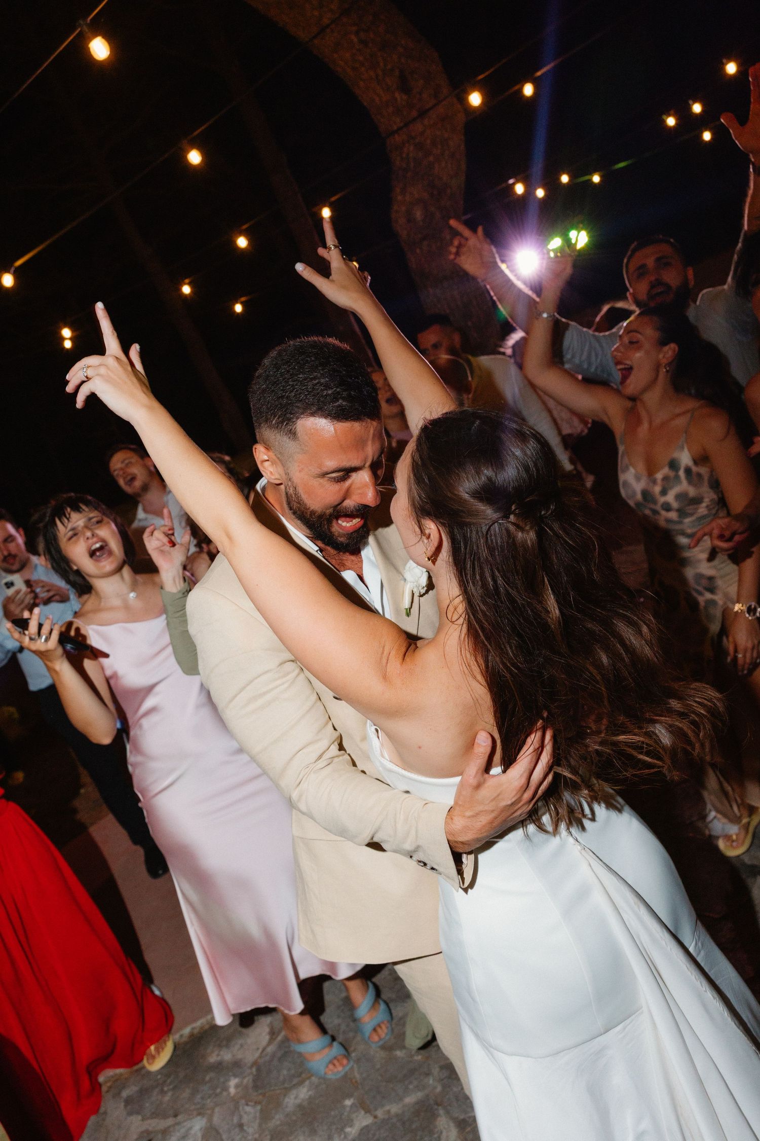 Bride and groom dancing joyfully surrounded by celebrating guests under string lights at night.