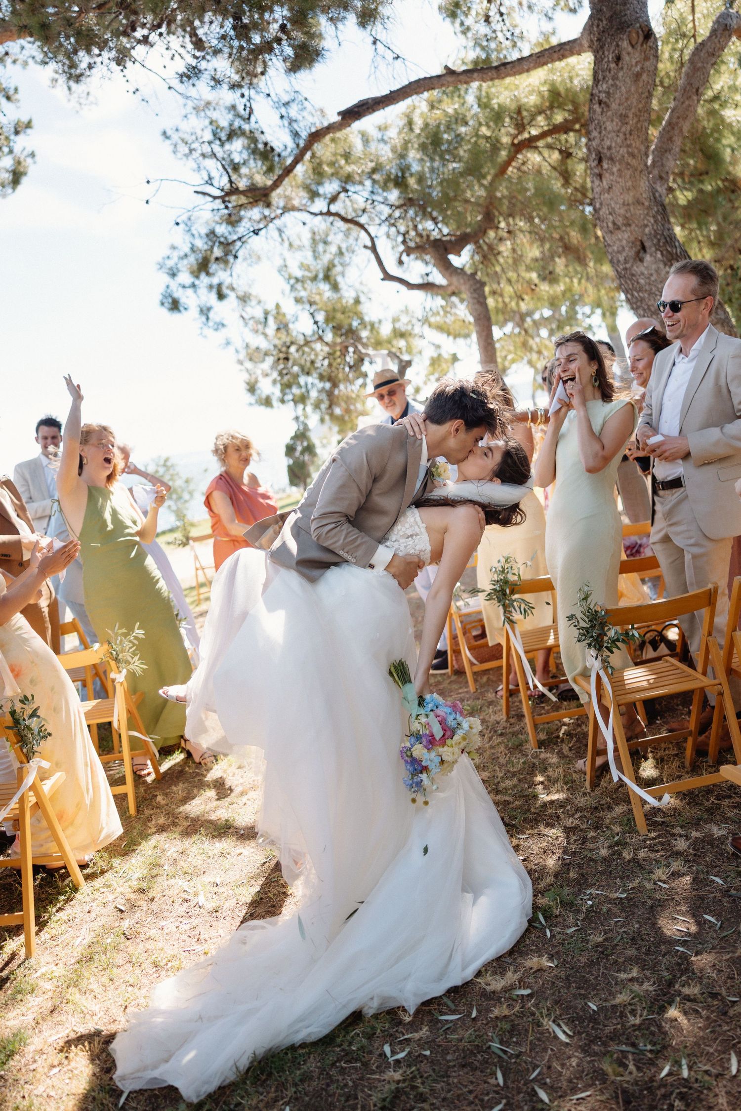 Bride and groom share a romantic dip kiss at outdoor wedding ceremony while guests celebrate among trees.