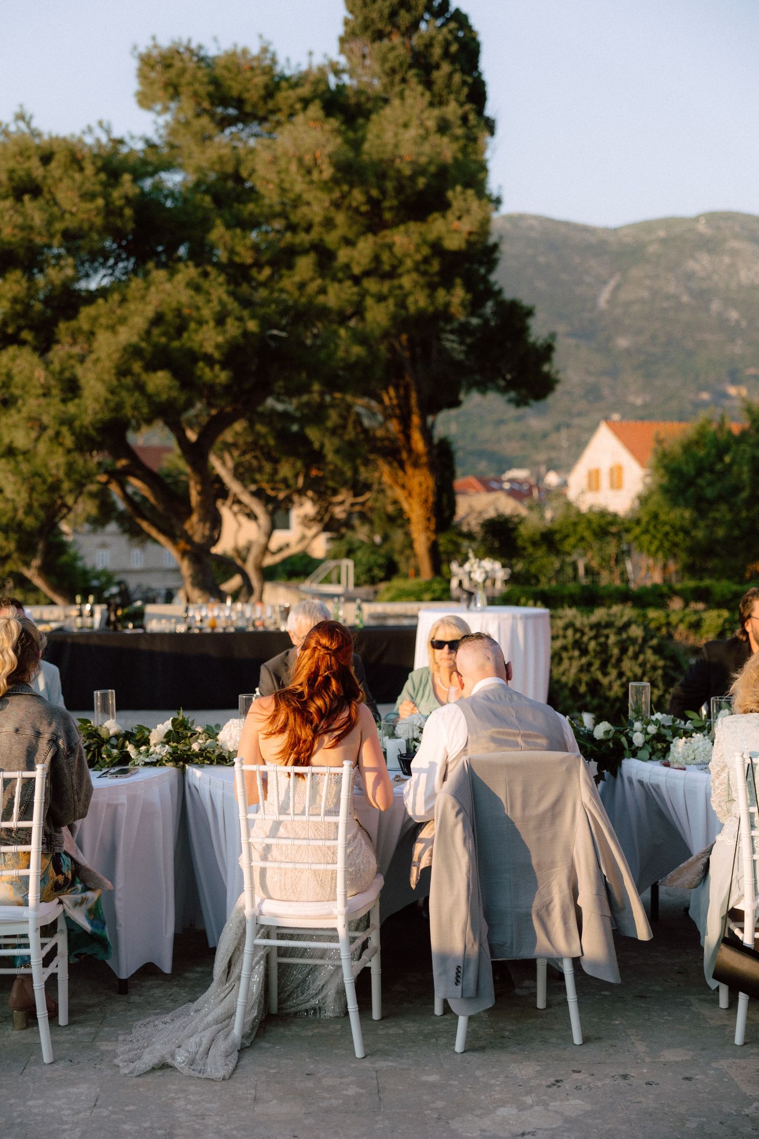 Wedding guests seated at long tables under trees during golden hour with mountain views.