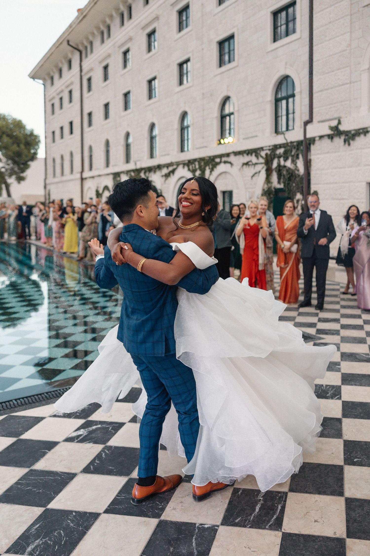 Groom lifts beaming bride in white gown on checkered dance floor at elegant outdoor wedding reception.
