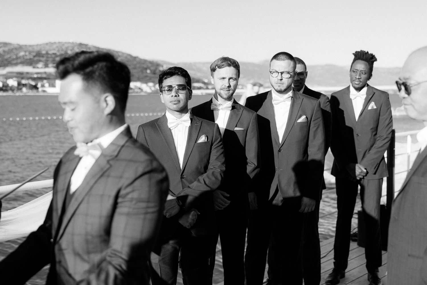 Groomsmen in dark suits stand by waterfront watching groom in foreground.