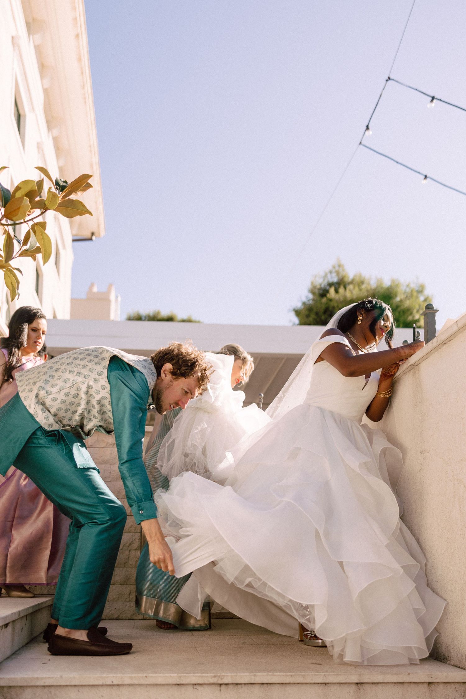 Groom in teal suit helps bride with wedding dress on outdoor steps during sunny ceremony.