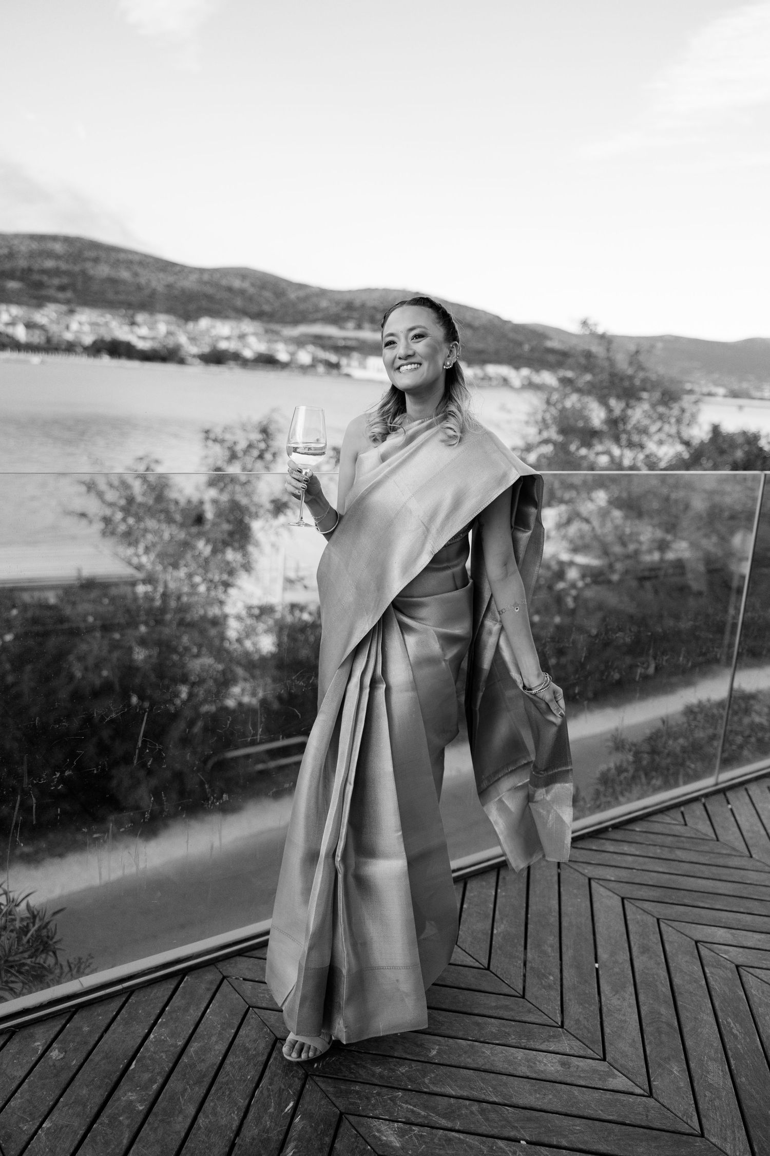 Woman in elegant sari holds wine glass on balcony with scenic waterfront view.