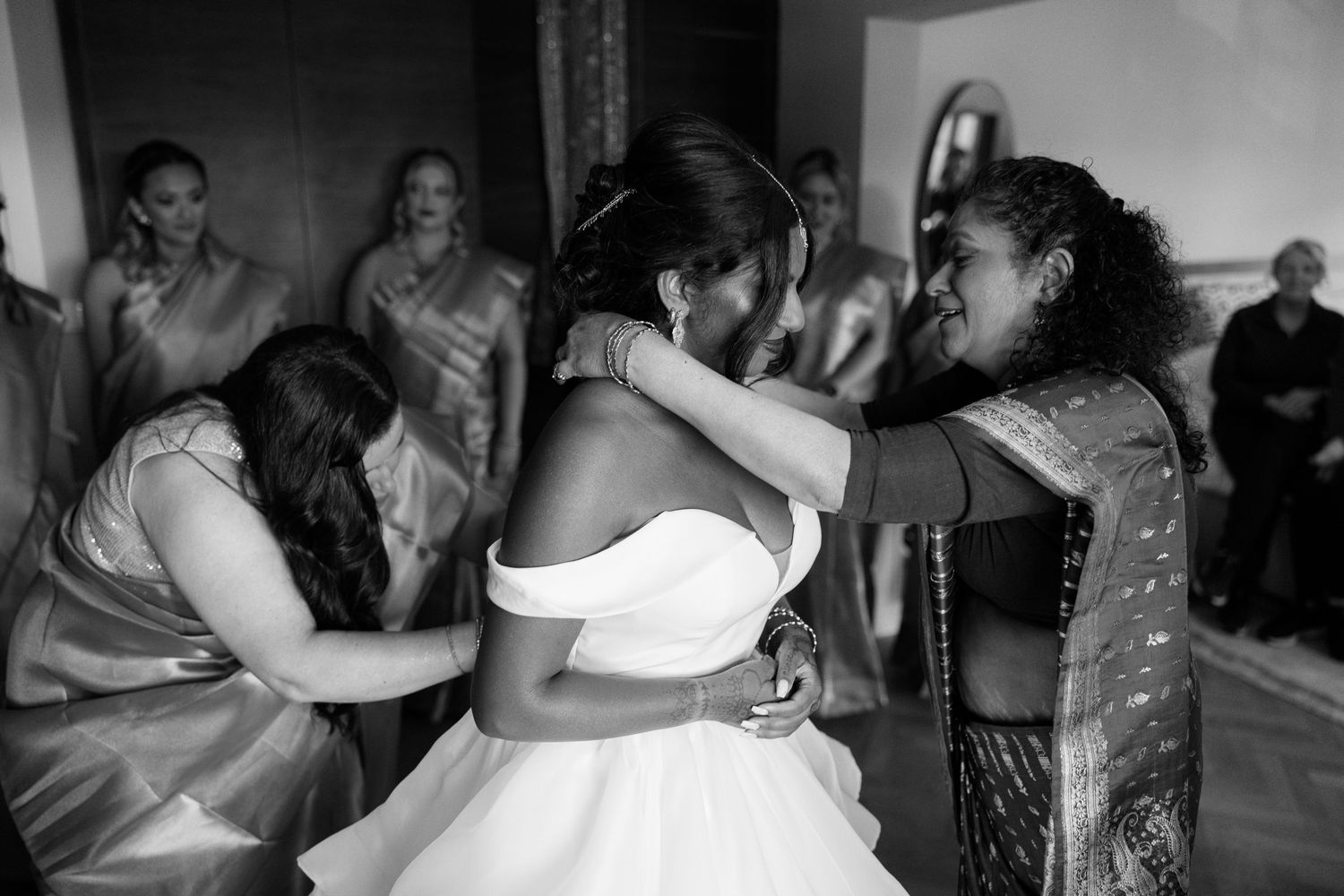 Bridesmaids helping bride into her white off-shoulder wedding gown in emotional getting-ready moment.