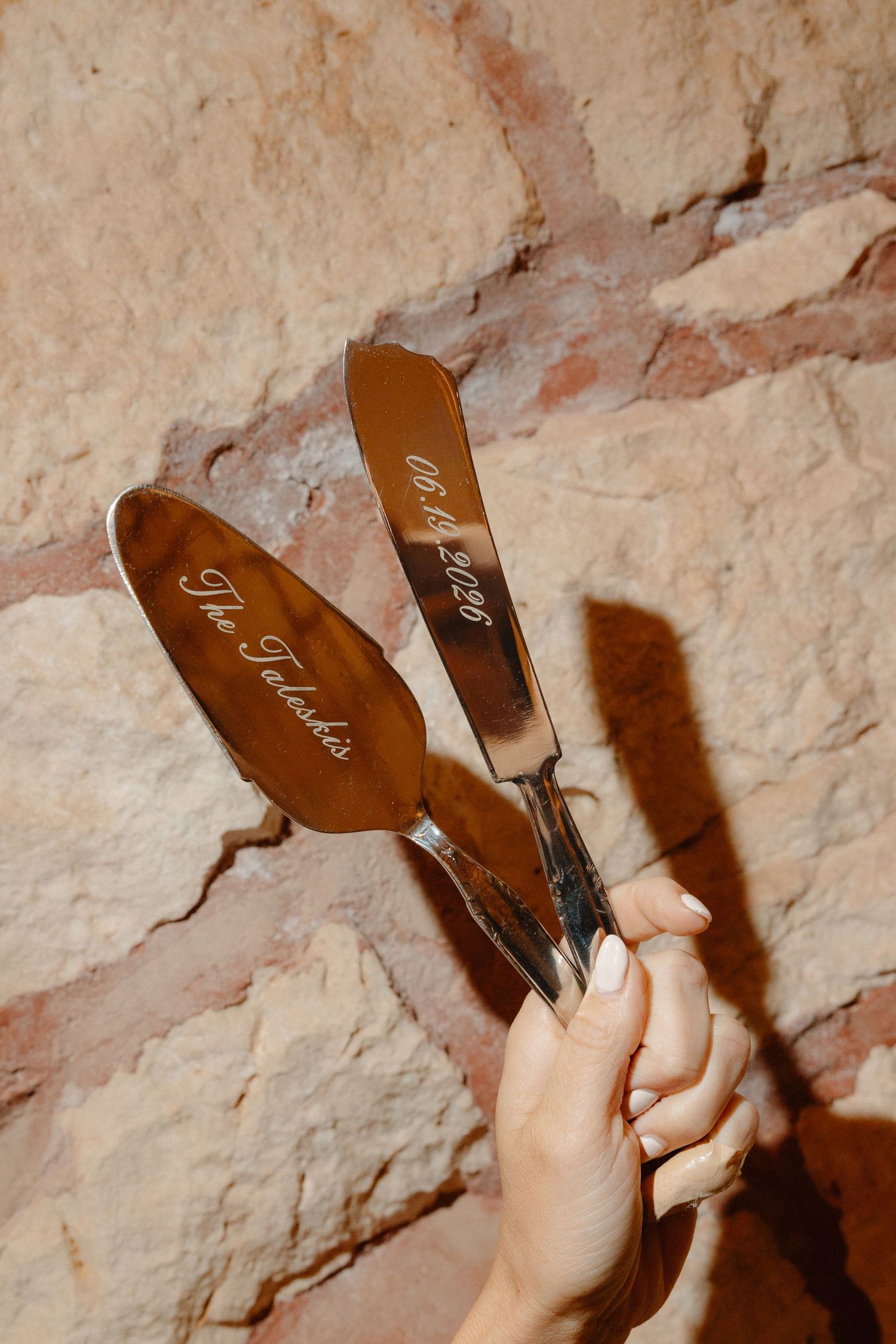 Hand holding personalized engraved wedding cake server set against rustic stone wall.