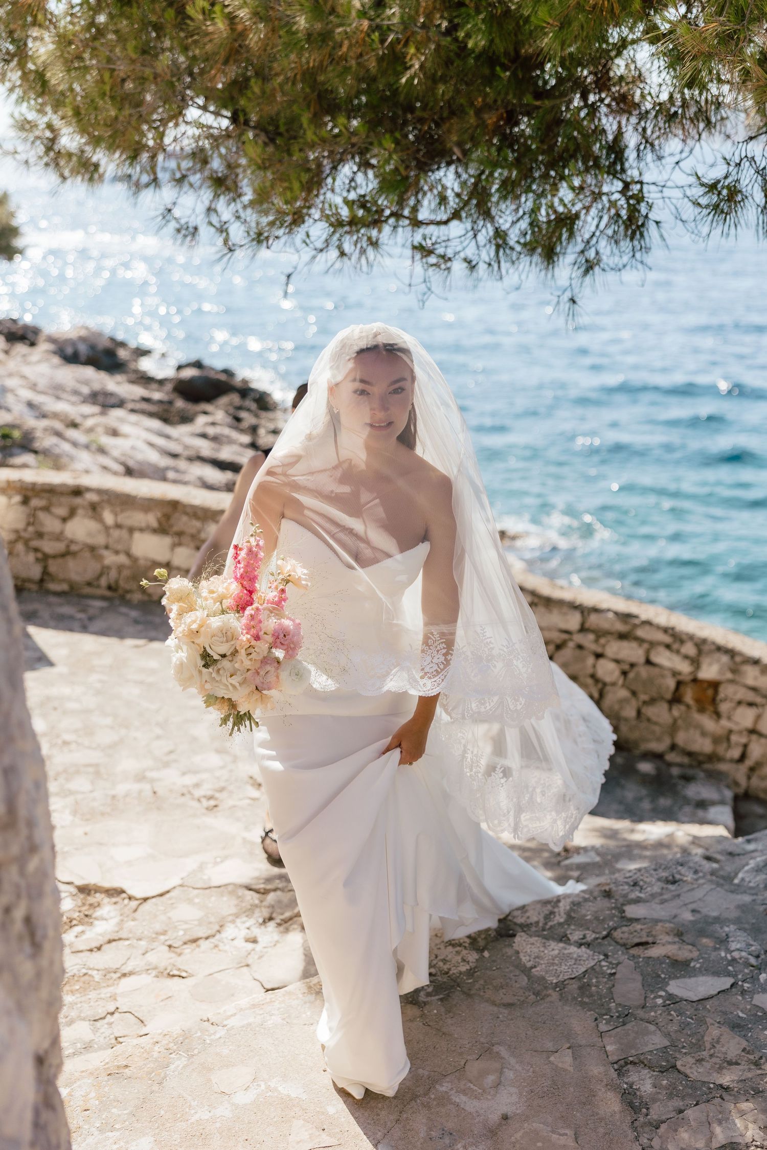 Bride in white gown and veil holds floral bouquet on stone path by sparkling blue sea.