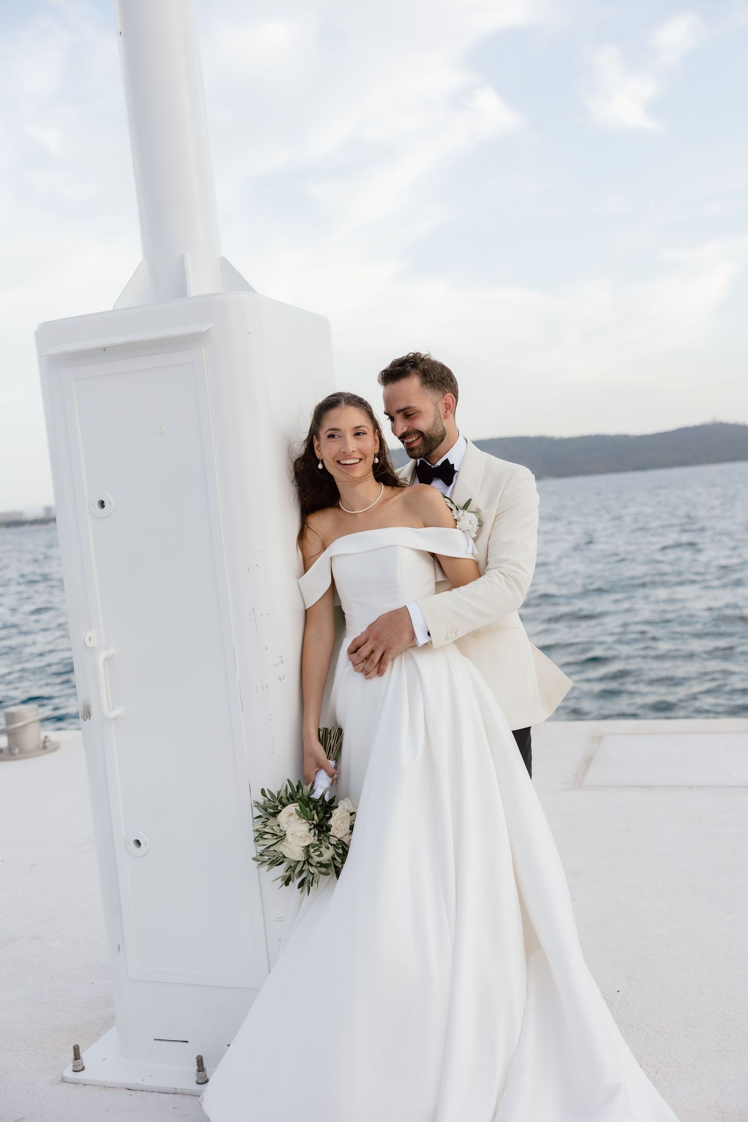 Bride and groom pose by white pillar overlooking ocean, she holds bouquet in off-shoulder gown, he wears cream tuxedo.