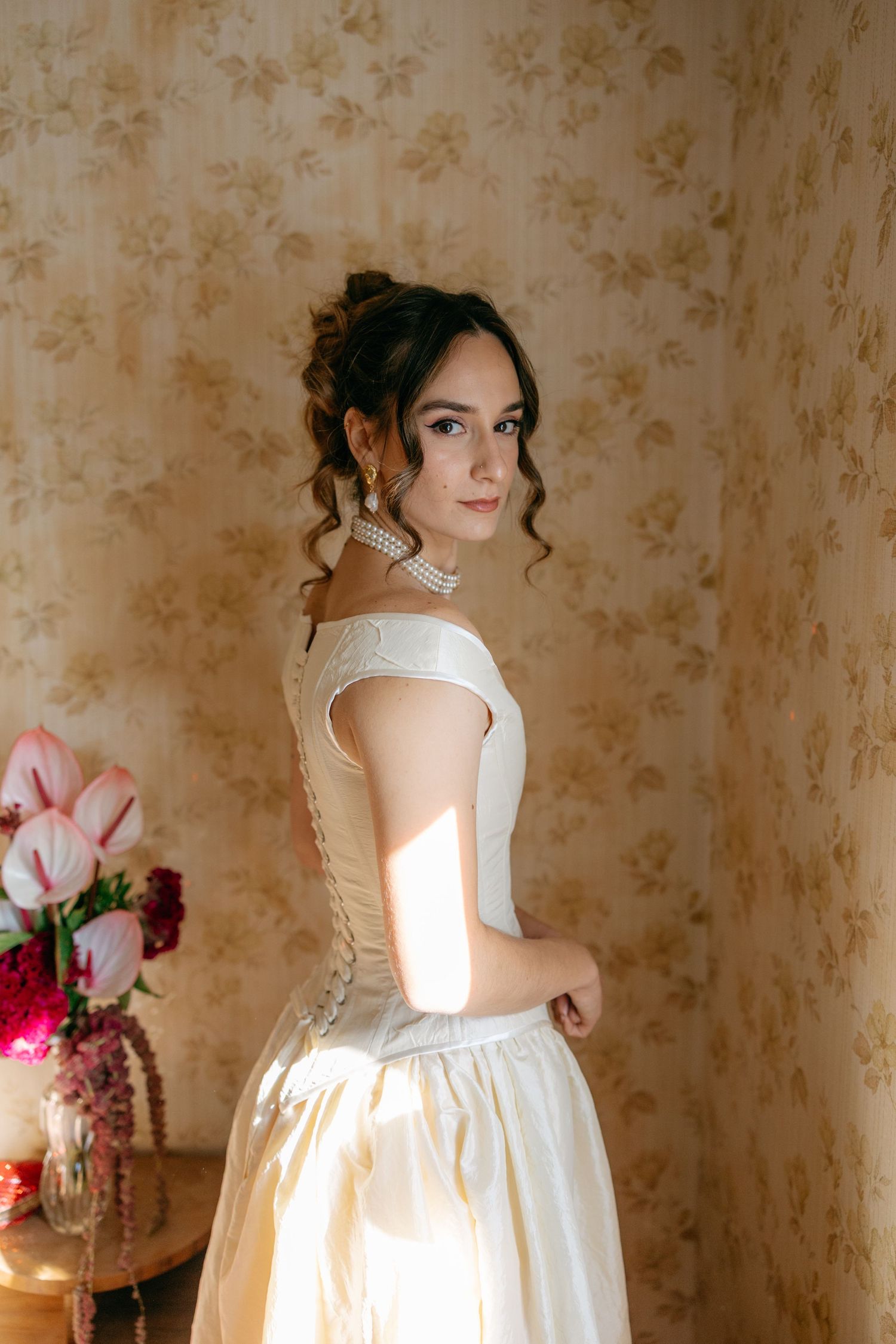 A bride in an elegant off-shoulder white wedding gown poses against a floral wallpaper background.